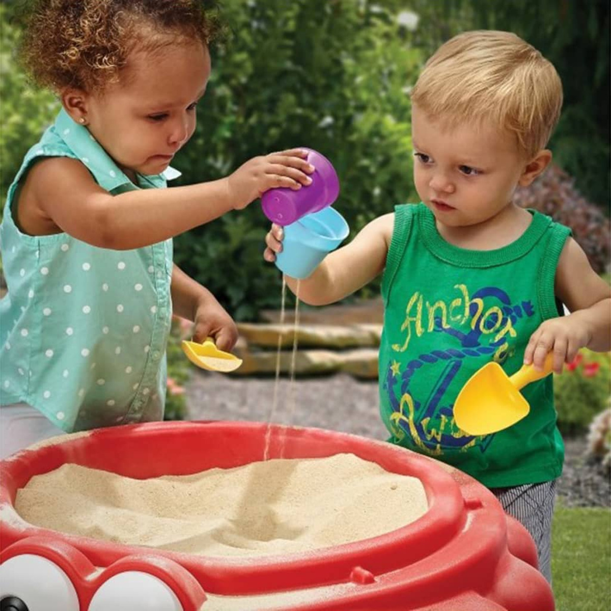 Zwei Kleinkinder spielen mit Sand und schütten ihn aus bunten Bechern in einen roten Krebssandkasten.