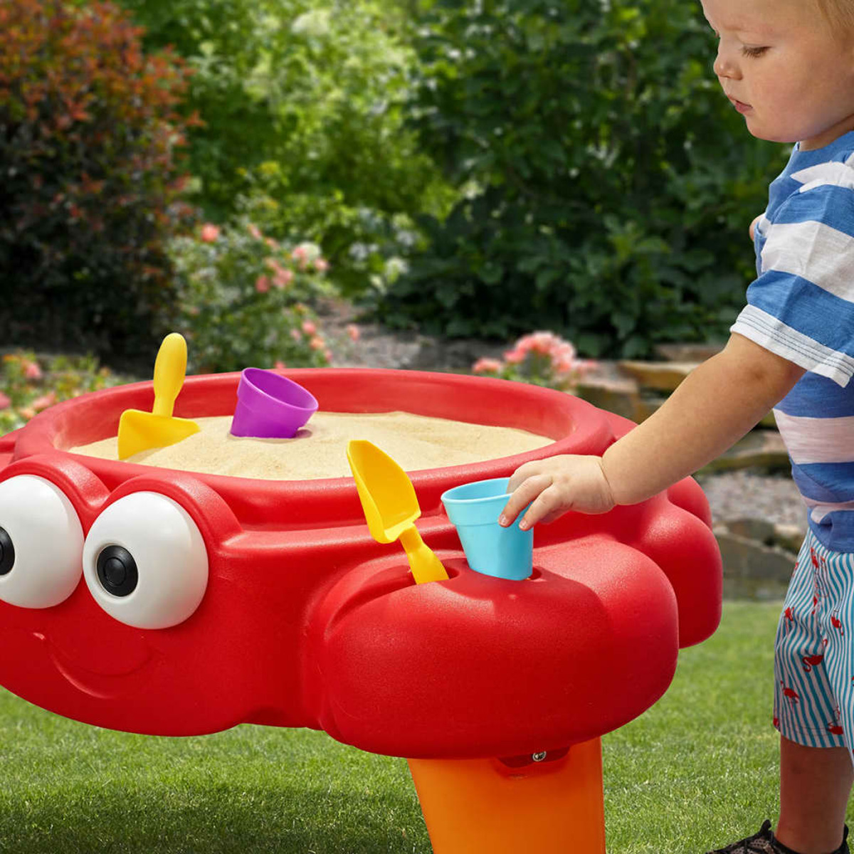 Ein Junge spielt an einem roten Krabben-Sandtisch im Garten mit Sand und bunten Förmchen.
