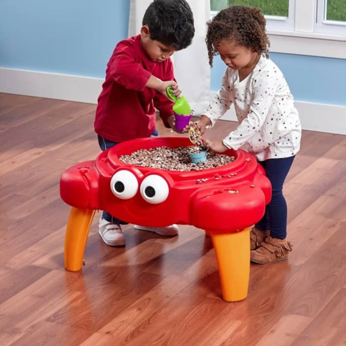 Zwei Kinder spielen an einem roten Krabben-Sand- und Wassertisch mit Muscheln.