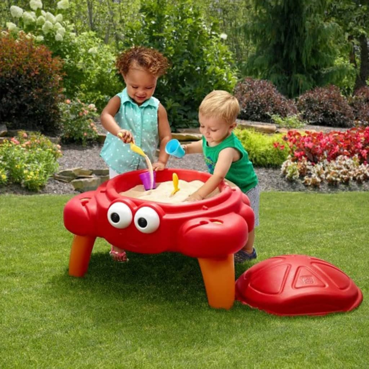 Zwei Kleinkinder spielen im Garten an einem roten Krabben-Sandkasten mit Sand und Förmchen.
