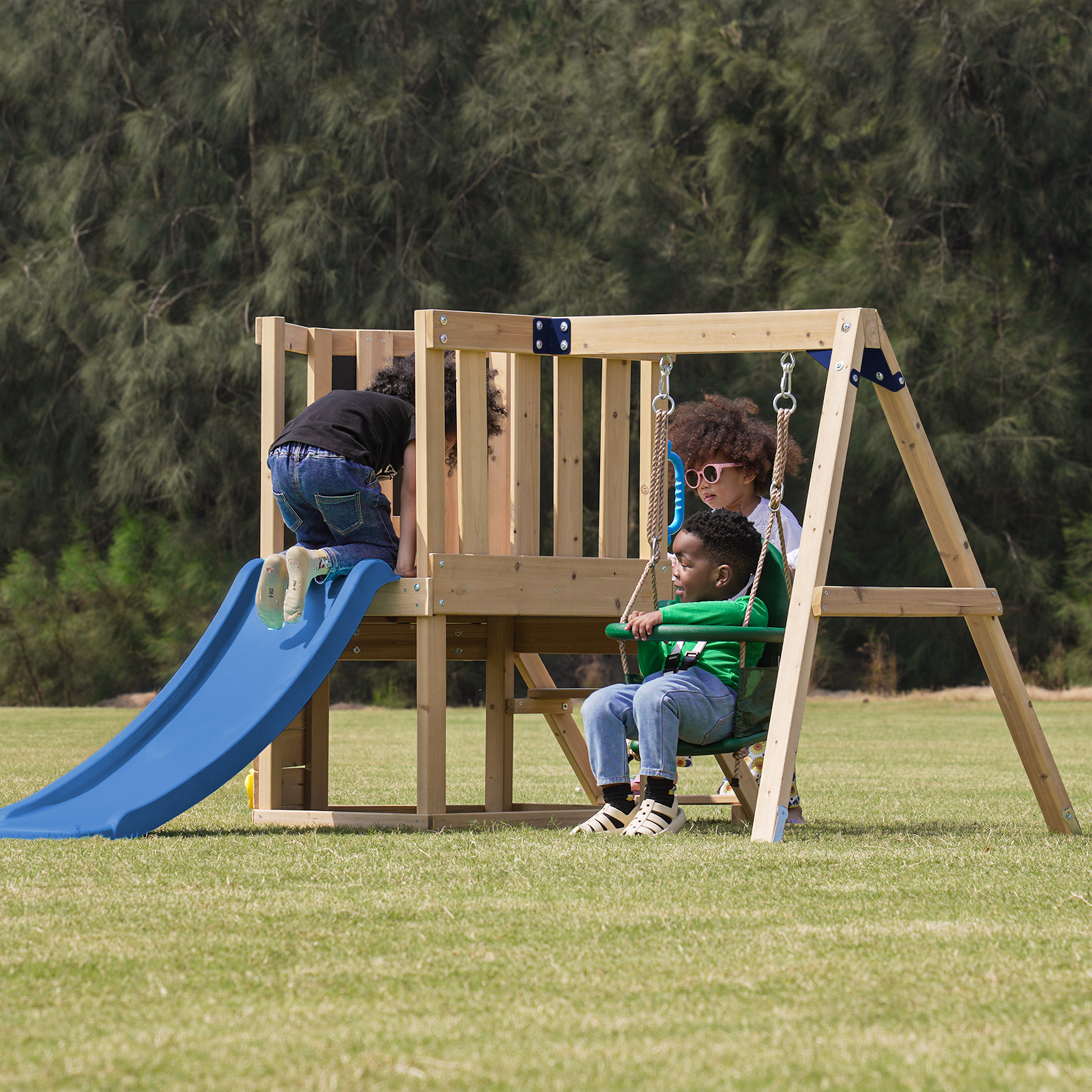 Drei Kinder spielen auf einem Holzspielplatz mit blauer Rutsche und grüner Schaukel auf einer Wiese.