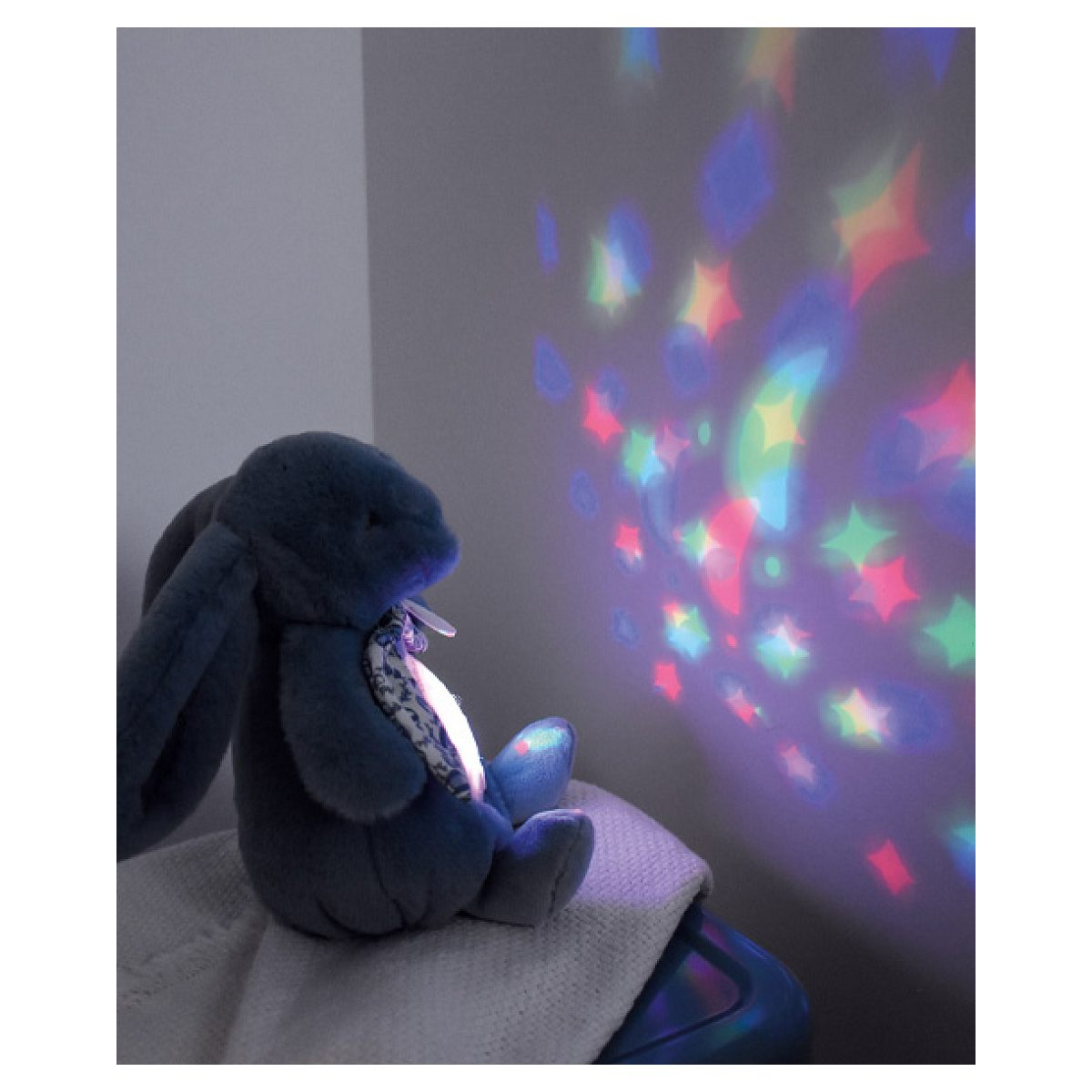 Ein blauer Plüschhase sitzt auf einem Handtuch und projiziert bunte Sterne und Lichter an die Wand.