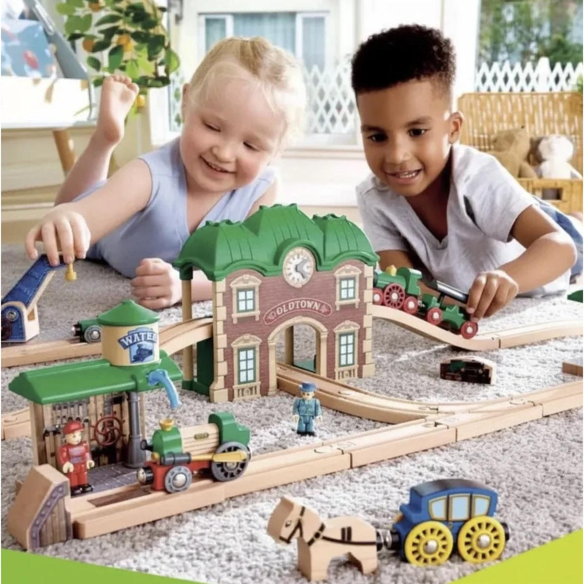 Zwei Kinder spielen glücklich auf dem Teppich mit einer Holzeisenbahn, Zügen und Gebäuden.