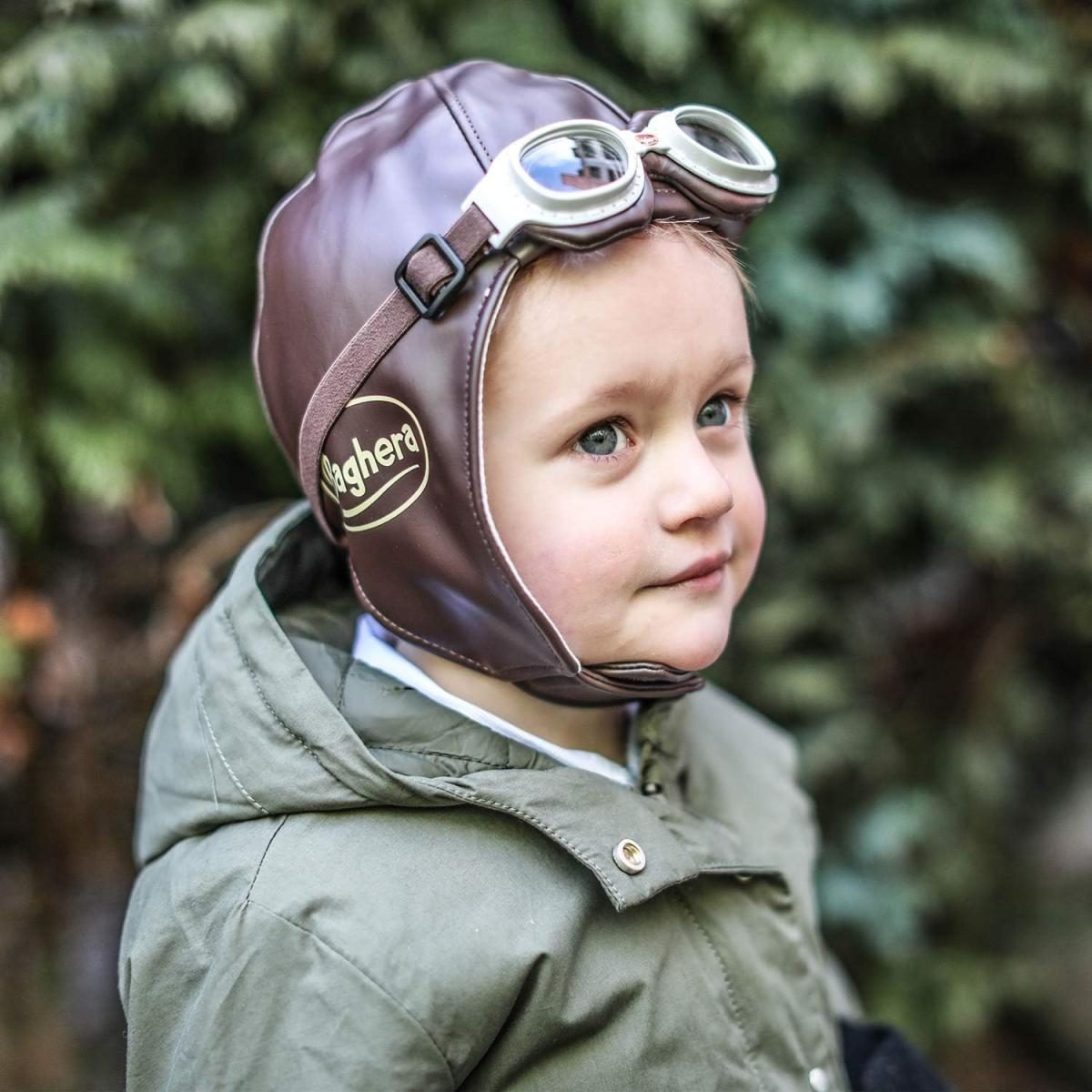 Ein kleines Kind in einer braunen Pilotenmütze und Brille mit einer grünen Jacke vor Bäumen.