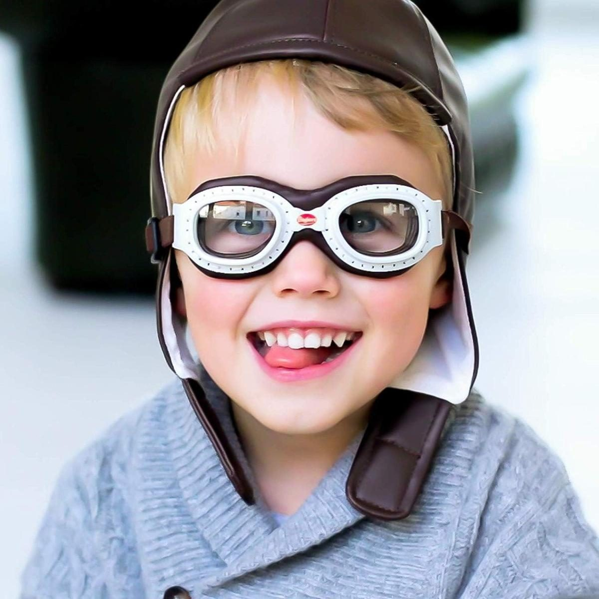 Ein blondes Kind mit Pilotenmütze und Schutzbrille lächelt und streckt die Zunge heraus.