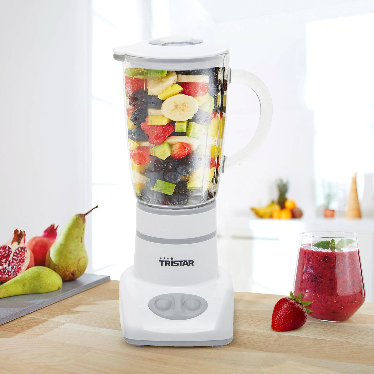 Un frullatore Tristar bianco pieno di frutta mista su un bancone di legno accanto a un bicchiere di smoothie e frutta.