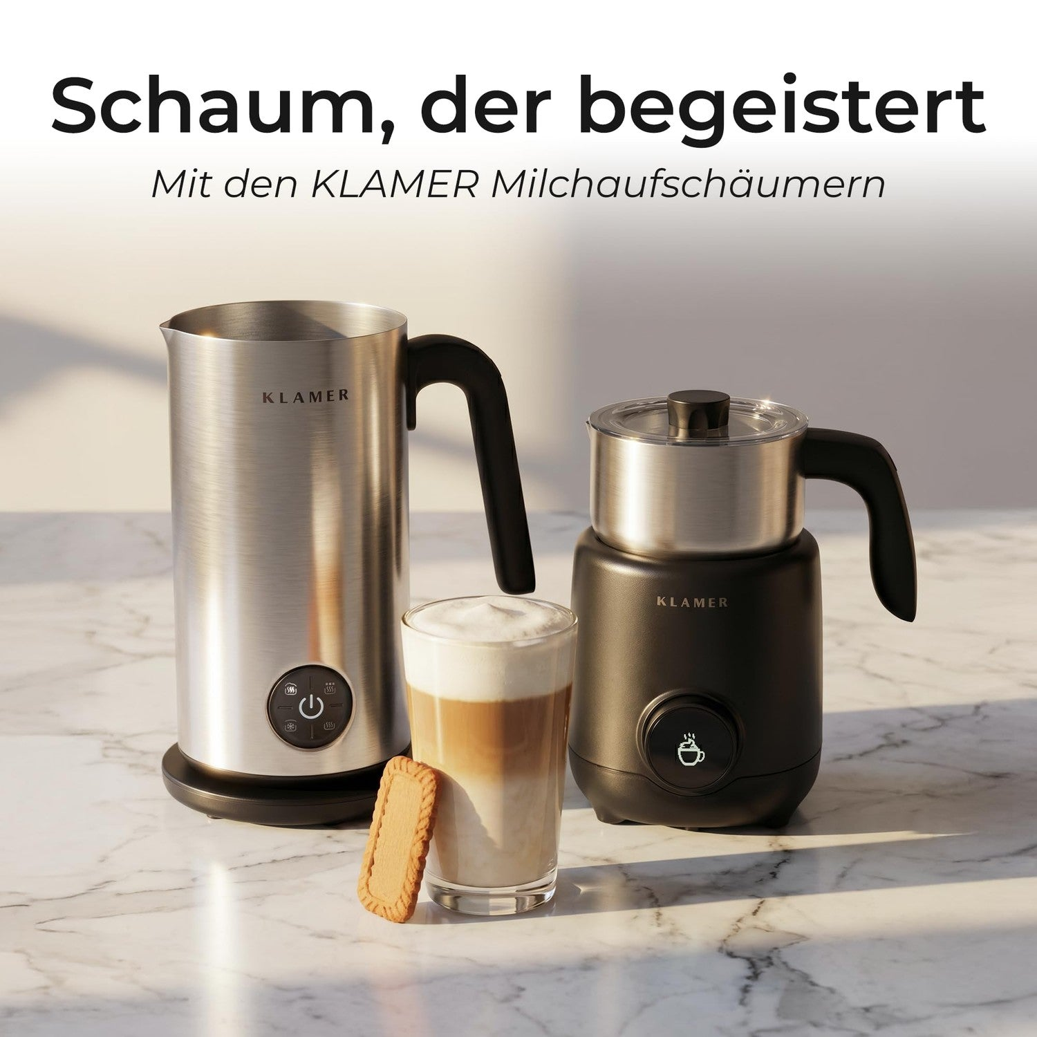 Zwei Klamer Milchaufschäumer, ein Latte Macchiato im Glas und ein Keks stehen auf einer Marmorarbeitsplatte.