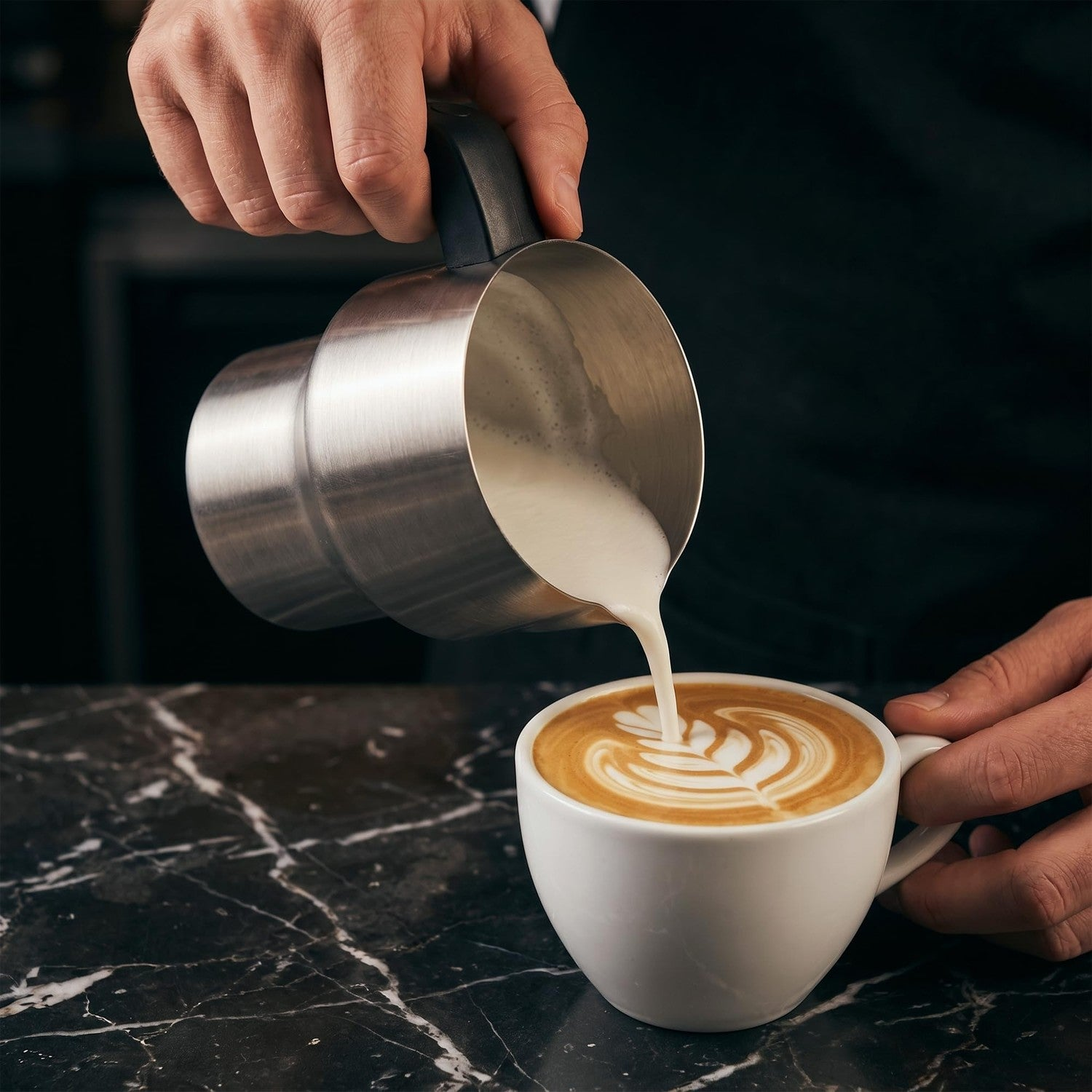 Ein Barista gießt Milch aus einer Kanne in eine weiße Tasse und kreiert Latte Art auf einer Marmortheke.