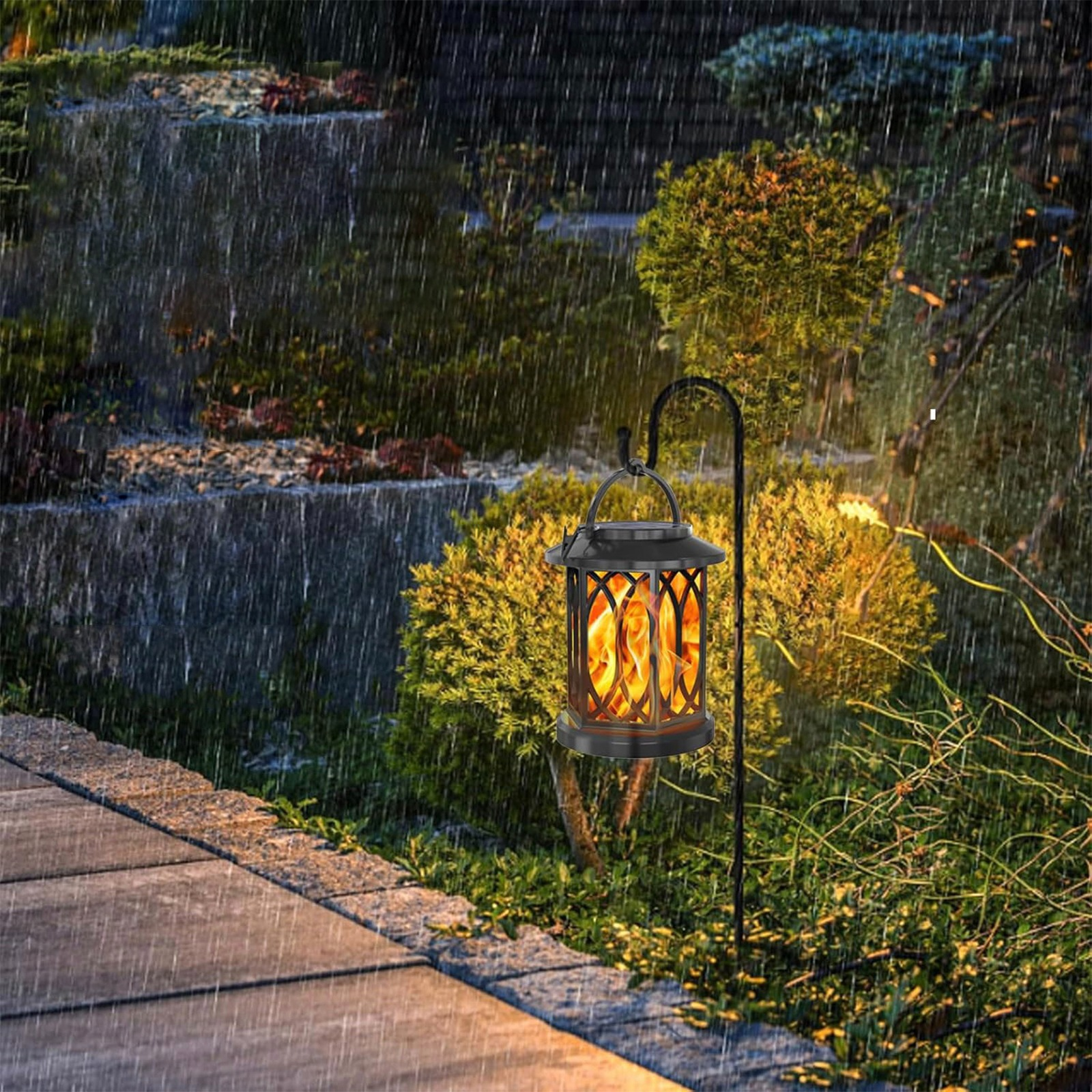 Eine schwarze Laterne mit Flammeneffekt hängt im Regen an einem Haken im Garten neben einem Weg.