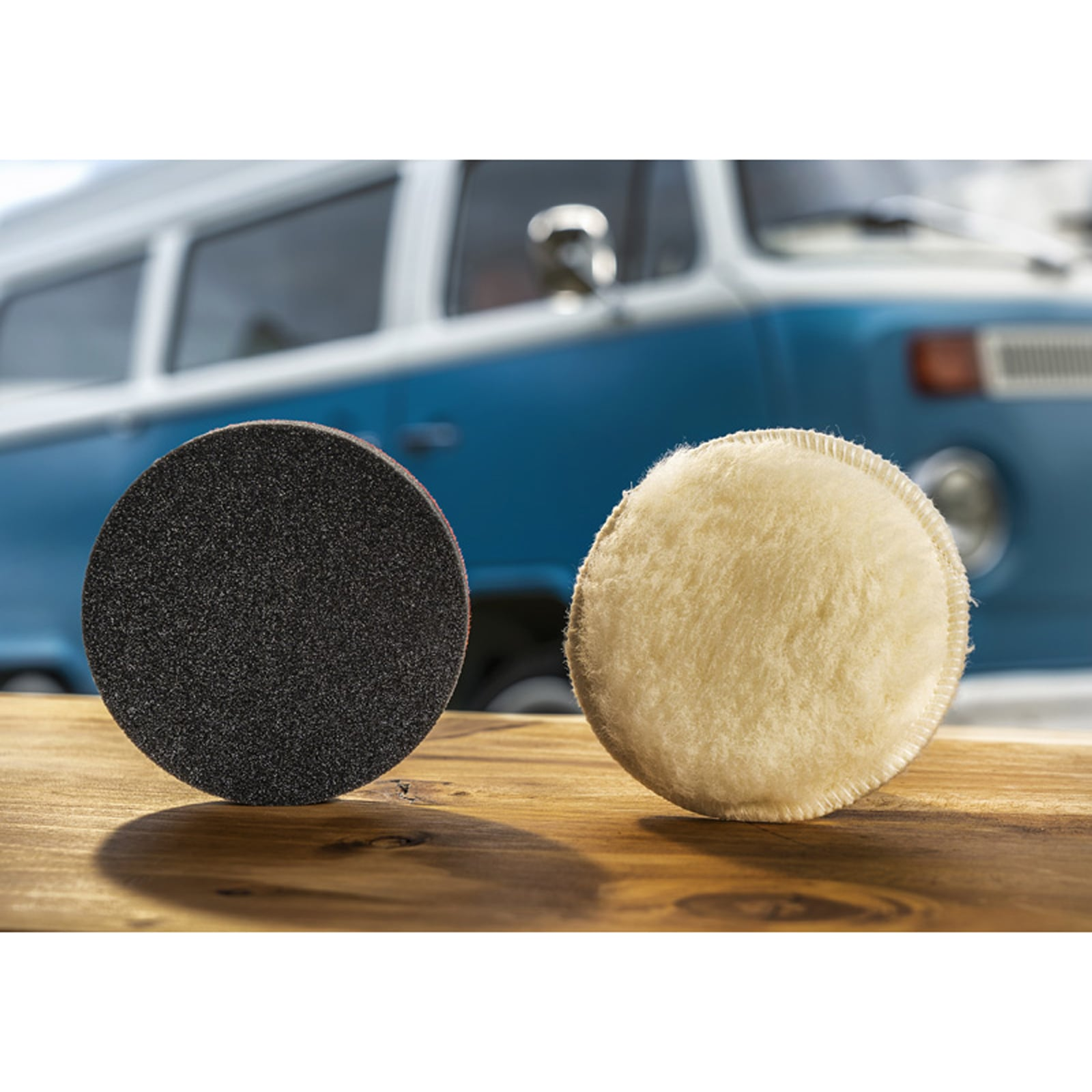 Eine schwarze Schleifscheibe und ein weißes Wollpad stehen auf einem Holzbrett vor einem blauen Auto.