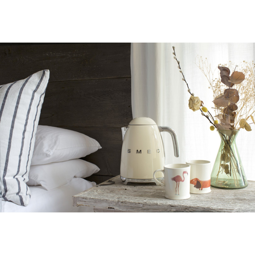 Ein beiger Wasserkocher, zwei Tassen und eine Blumenvase stehen auf einem Holztisch neben einem Bett.