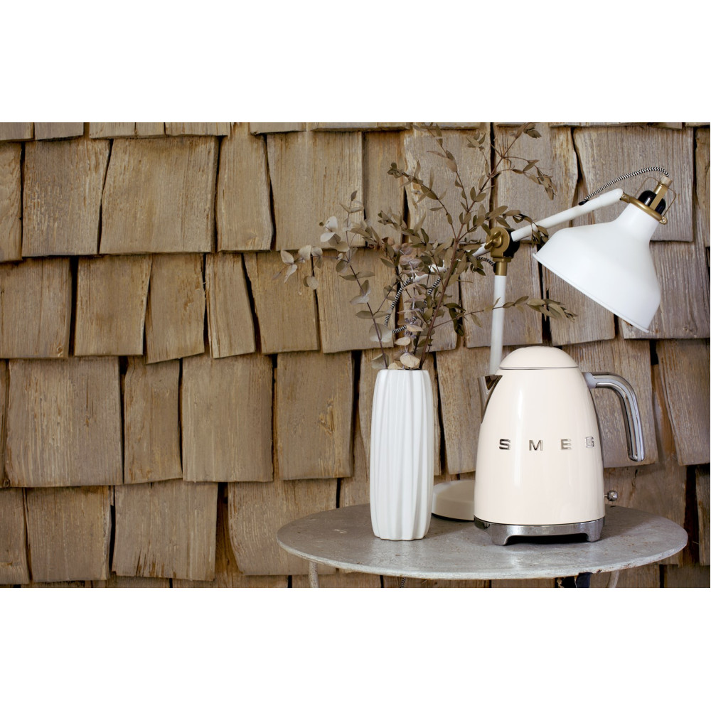 Cremefarbener Smeg-Wasserkocher, weiße Vase mit Zweigen und Lampe auf Metalltisch vor Holzwand.