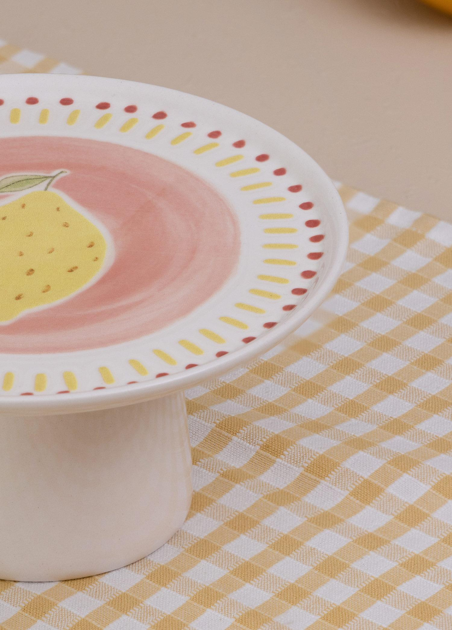 Pembe zemin üzerinde limon motifli beyaz bir kek standı, sarı-beyaz kareli bir masa örtüsü üzerinde duruyor.