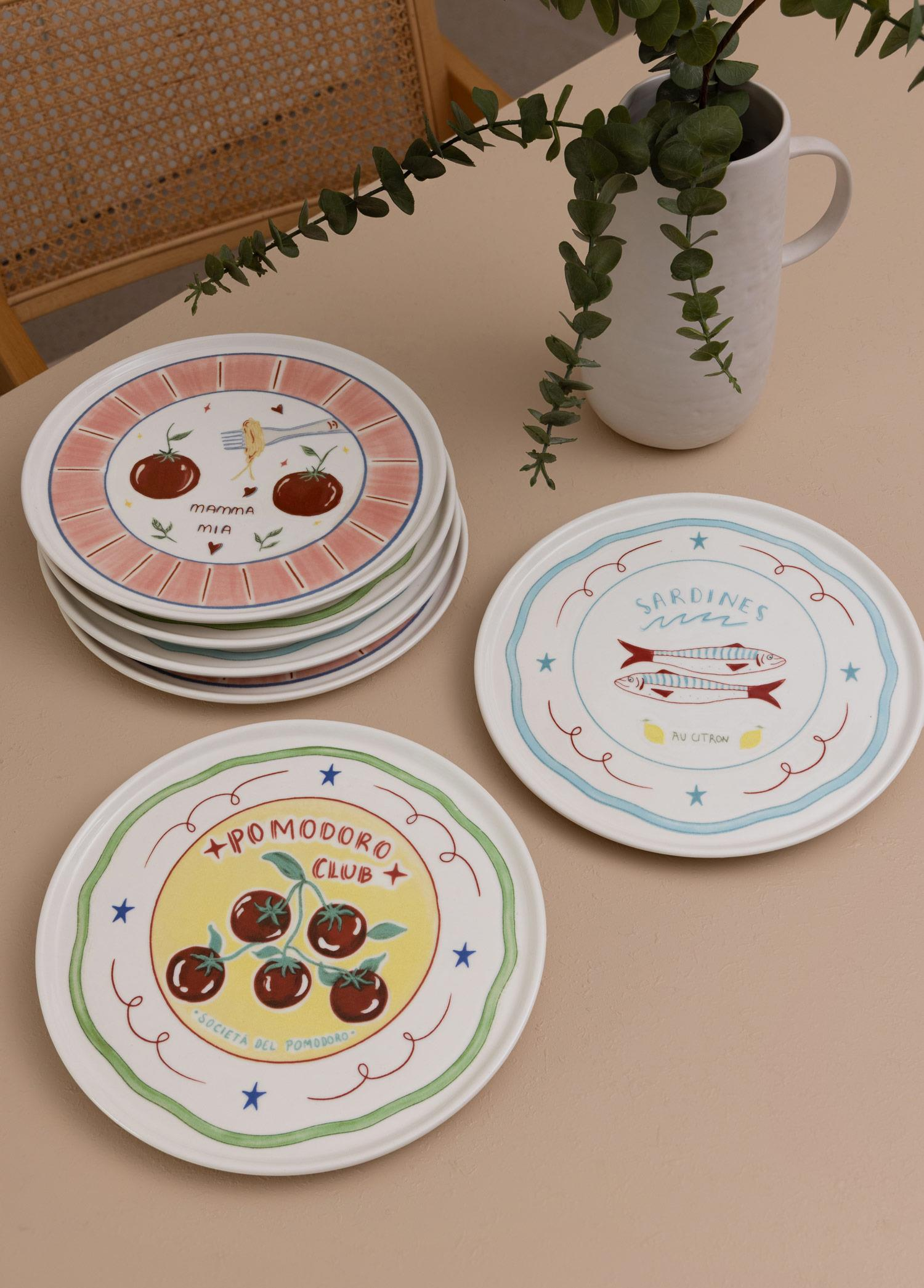 Masada yeşil dallı bir vazo yanında yemek motifli dekoratif tabaklar.