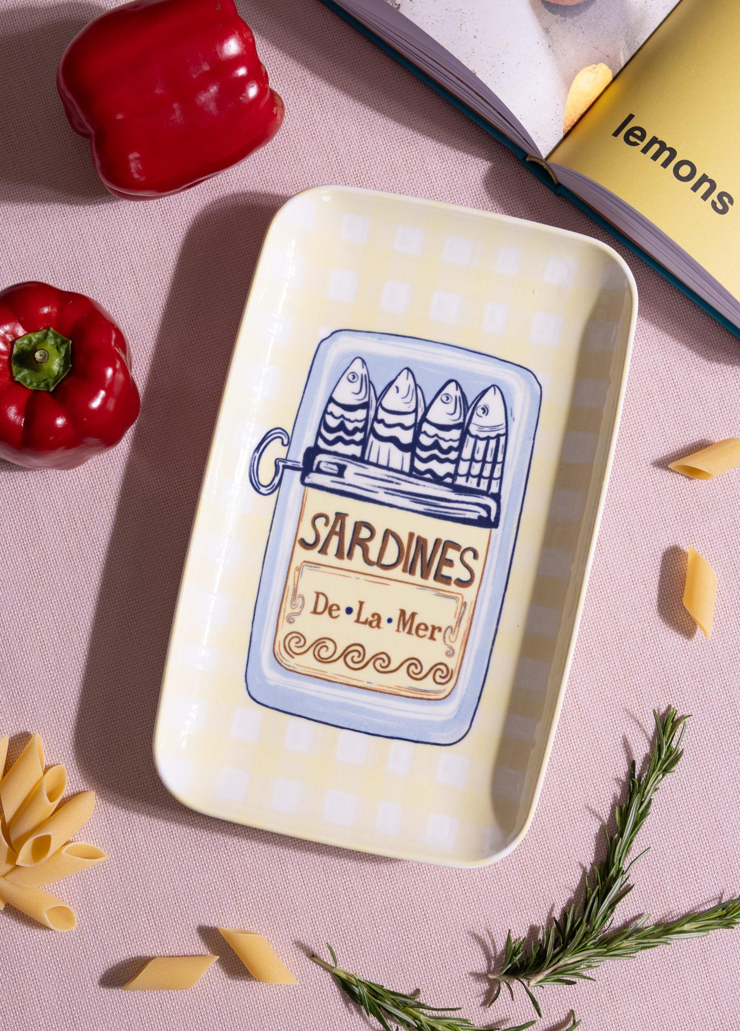 Pembe zemin üzerinde biber, makarna ve bir kitapla çevrili sardalya desenli sarı ekose tabak.