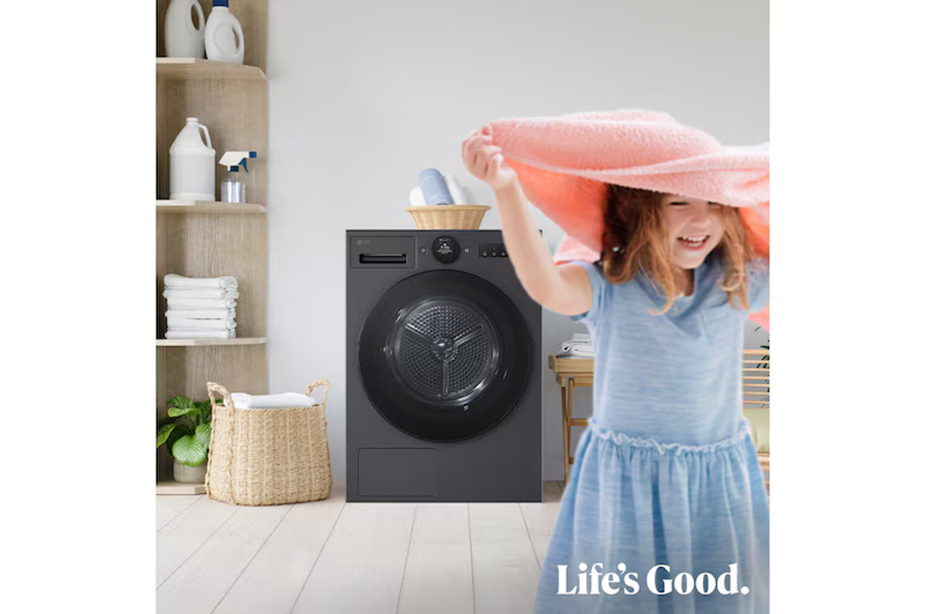 Una niña sonriente con vestido azul sostiene una toalla rosa frente a una lavadora oscura.