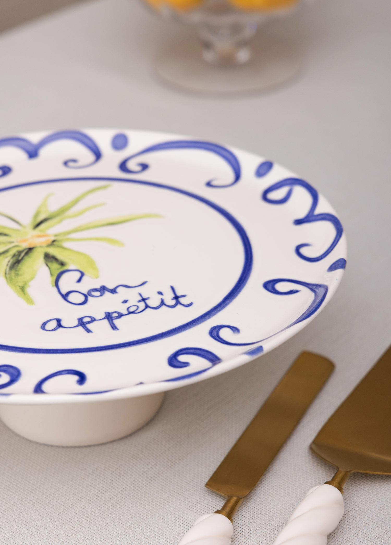 Mavi desenli beyaz bir kek standı, Bon Appetit yazısı ve yanında altın renkli çatal bıçak takımı.