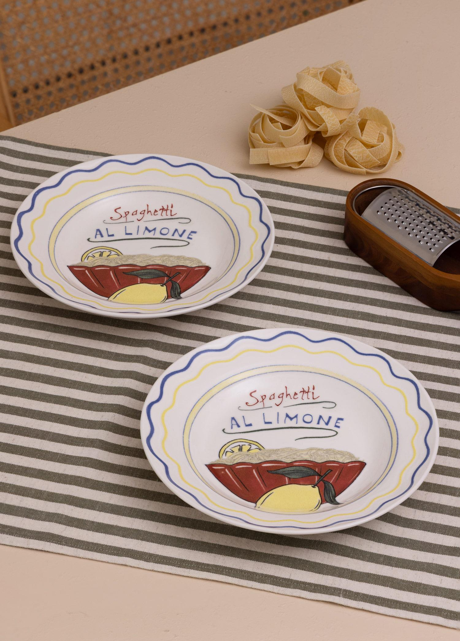 Spaghetti al Limone yazılı iki tabak, çiğ makarna yuvaları ve çizgili bir masa örtüsü üzerinde bir rende.