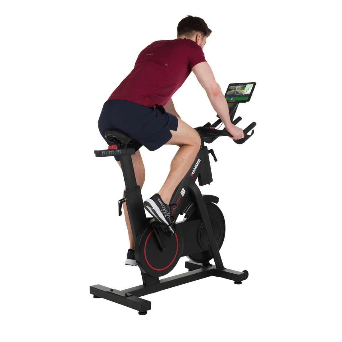 Ein Mann in rotem Shirt und blauen Shorts trainiert auf einem schwarzen Heimtrainer vor weißem Hintergrund.