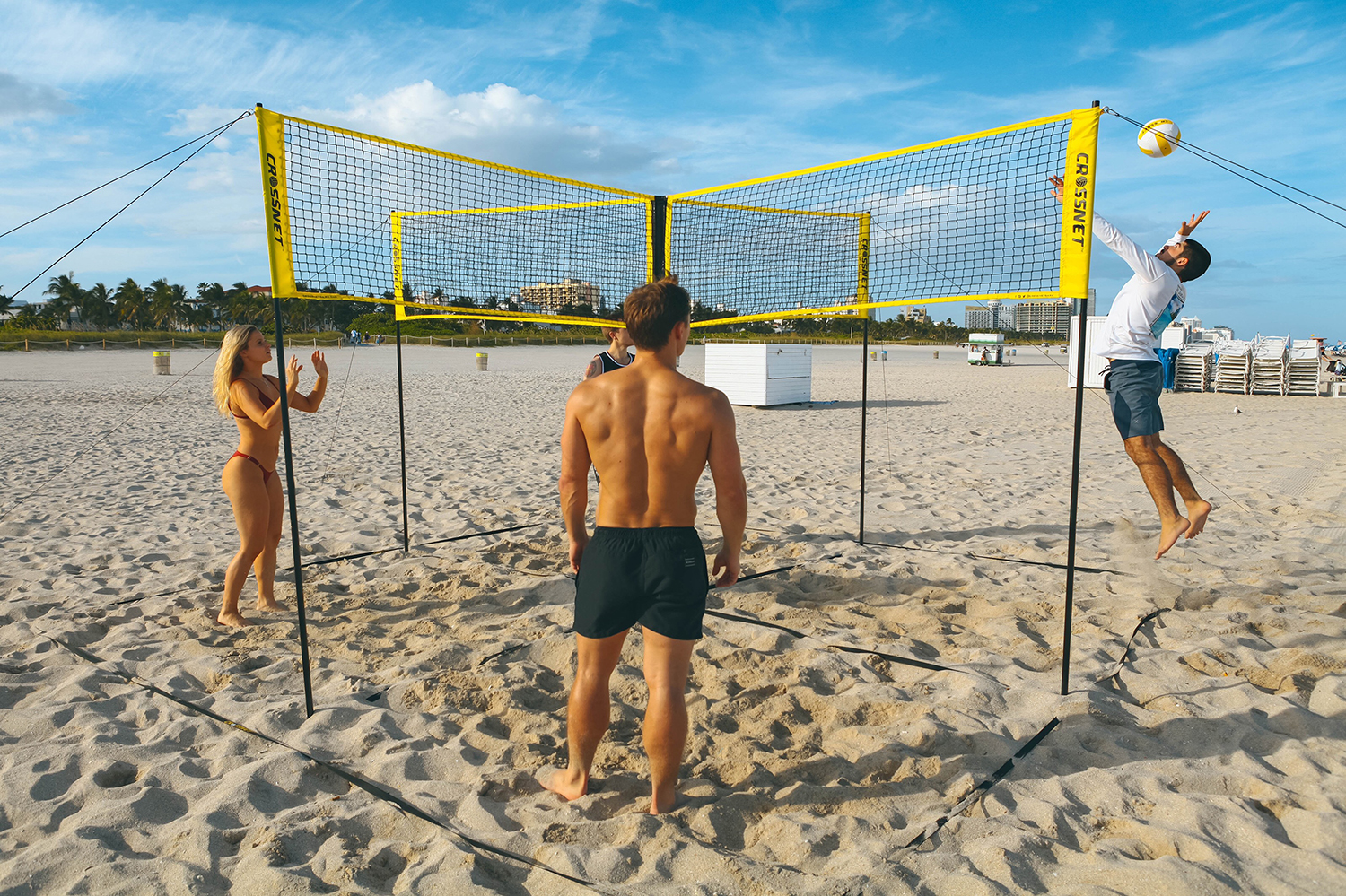 Vier Personen spielen Crossnet-Volleyball am sonnigen Strand unter blauem Himmel.