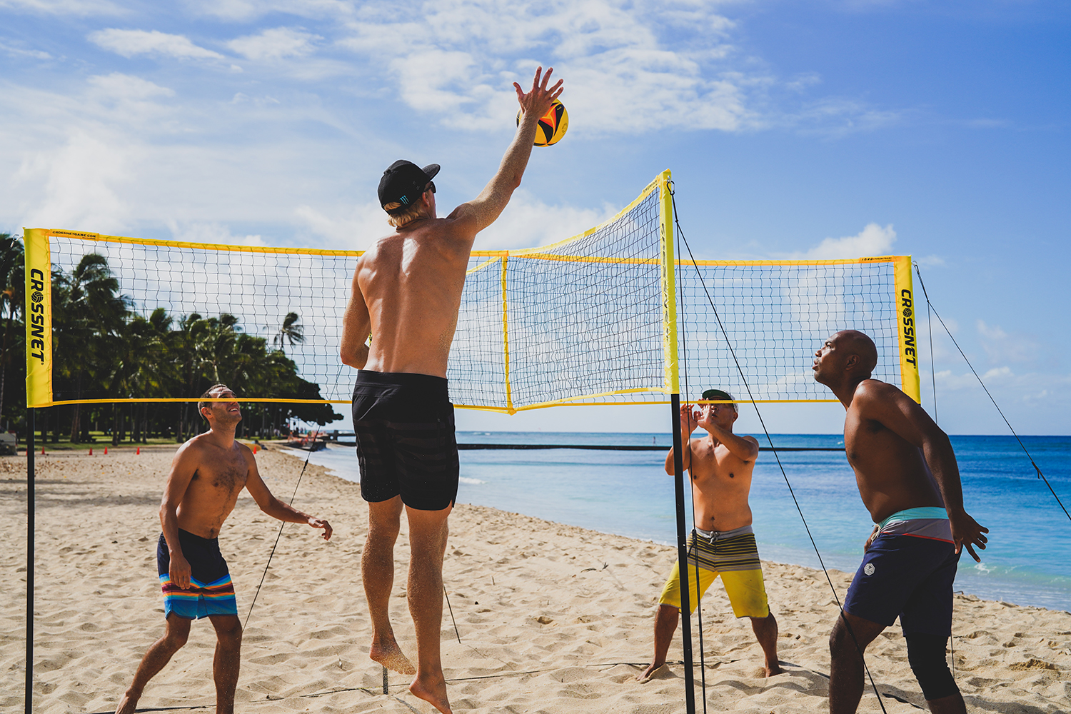 Vier Männer spielen Beachvolleyball an einem sonnigen Tropenstrand mit blauem Ozean im Hintergrund.