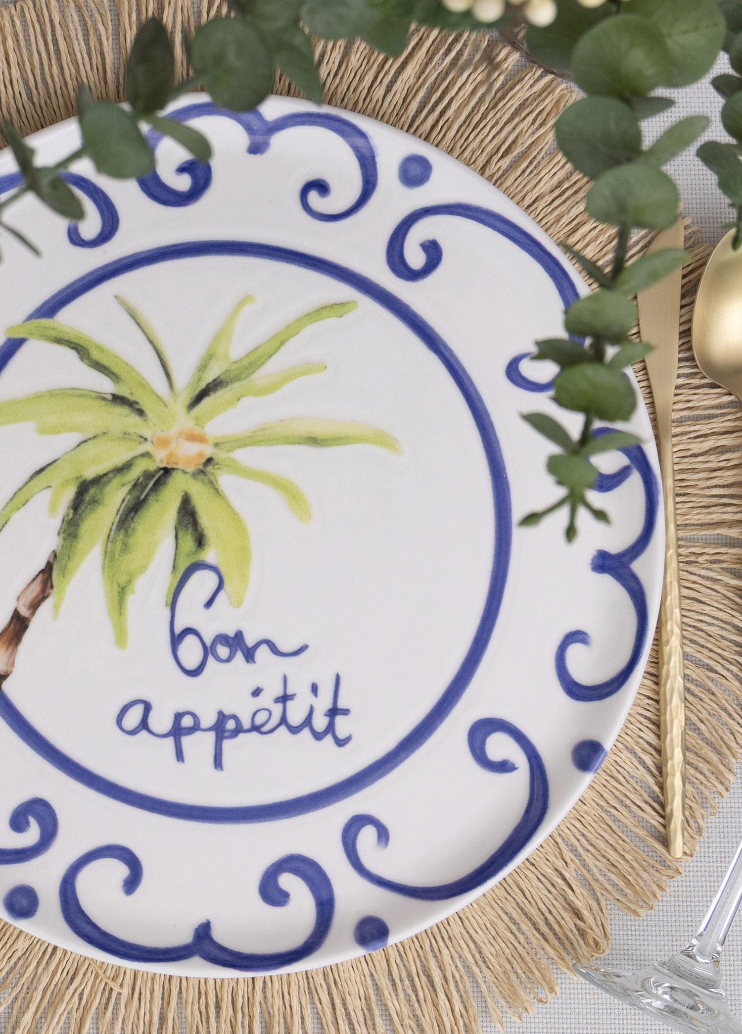 Mavi desenli, palmiye ağaçlı ve Bon Appetit yazılı beyaz bir tabak, hasır supla üzerinde çatal bıçakla.