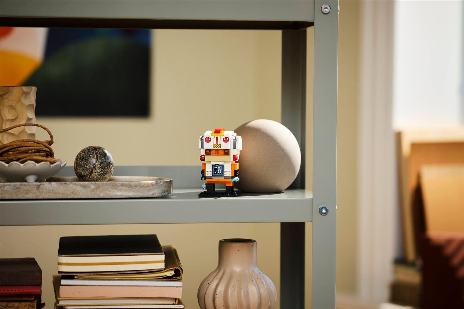 Eine kleine Roboterfigur aus Bausteinen steht auf einem grauen Regal, umgeben von Vasen und Büchern.