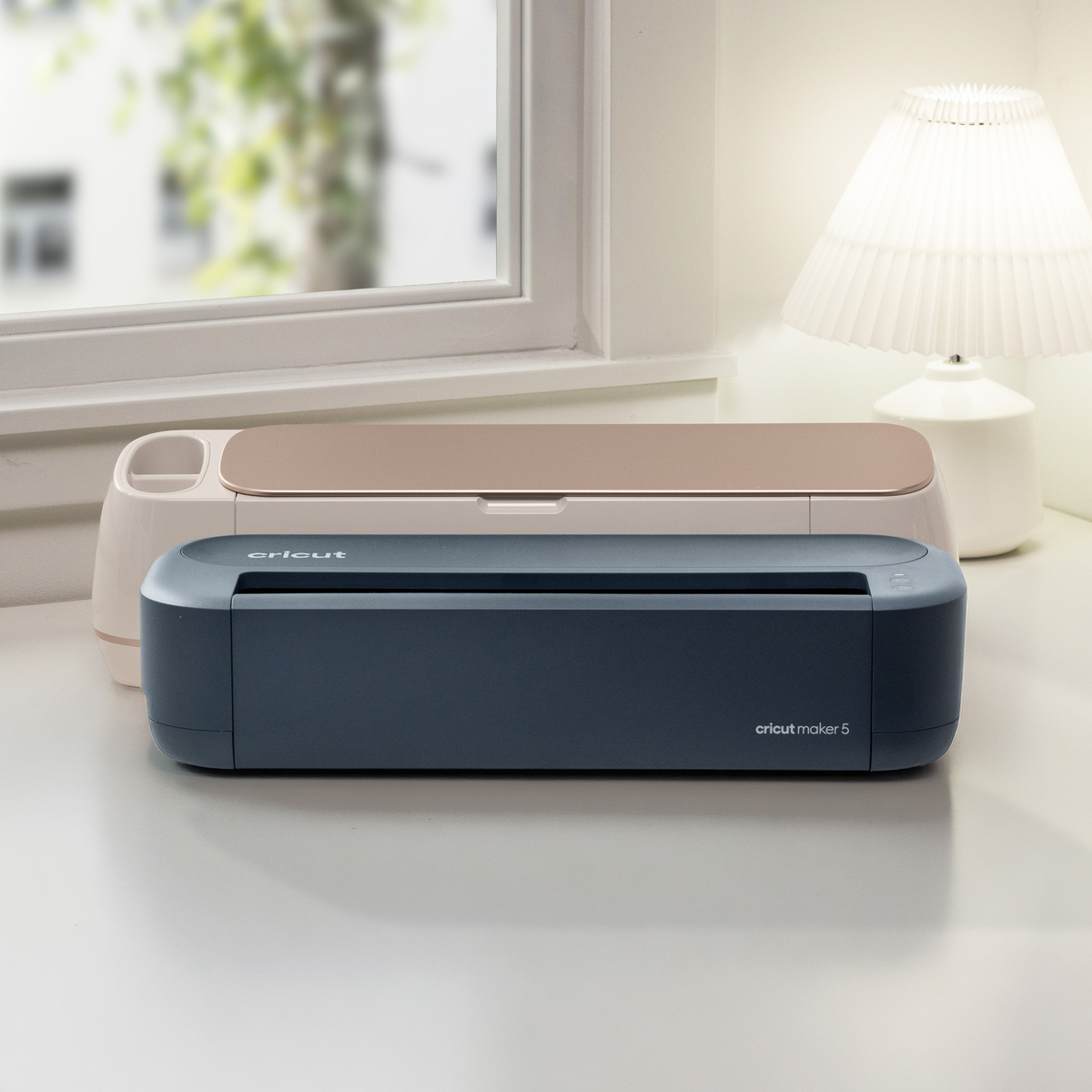 Dos máquinas Cricut, en beige y azul, están en una mesa junto a una ventana al lado de una lámpara.