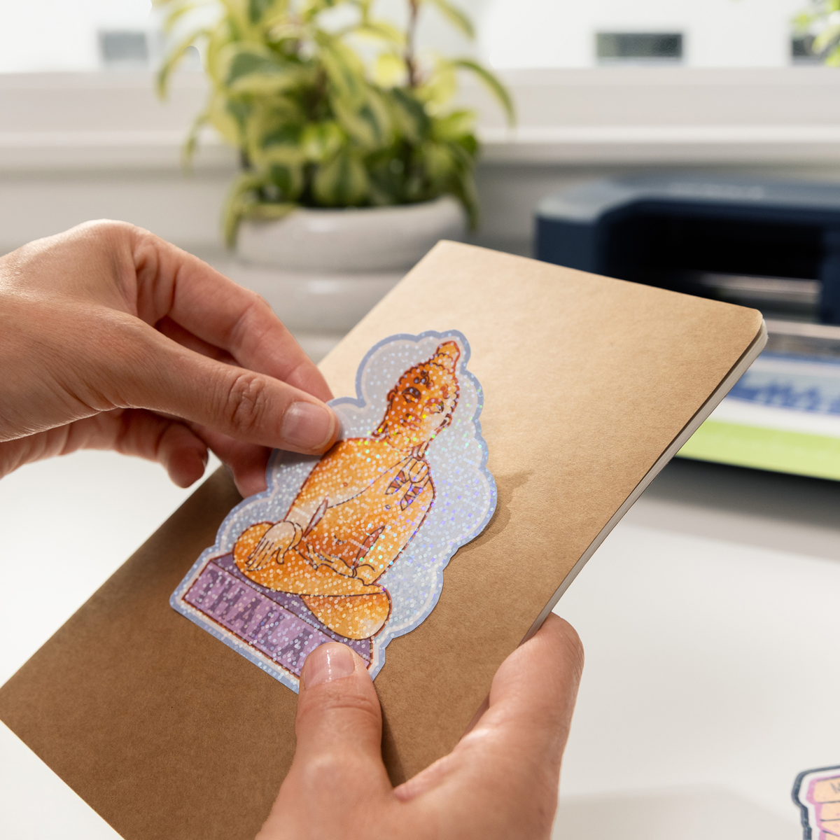 Persona colocando un adhesivo brillante de Buda en un cuaderno marrón.