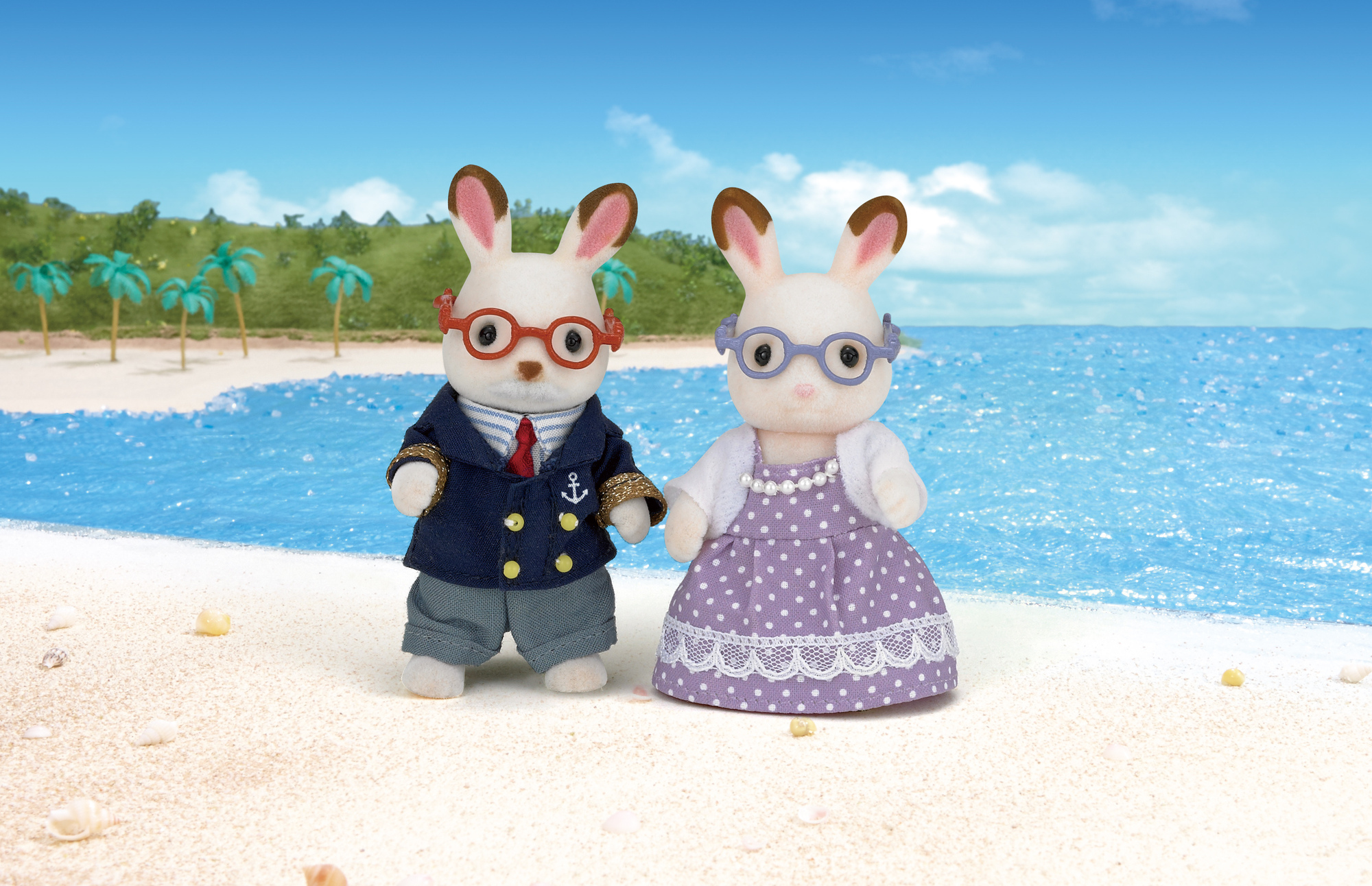 Zwei Plueschhasen in Kleidung stehen an einem sonnigen Sandstrand vor dem blauen Meer.