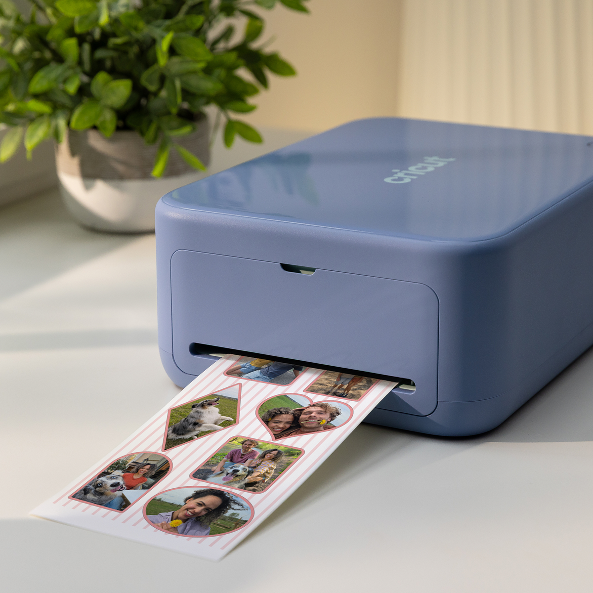 Una impresora Cricut azul en una mesa blanca imprime una hoja de pegatinas con fotos de personas y perros.