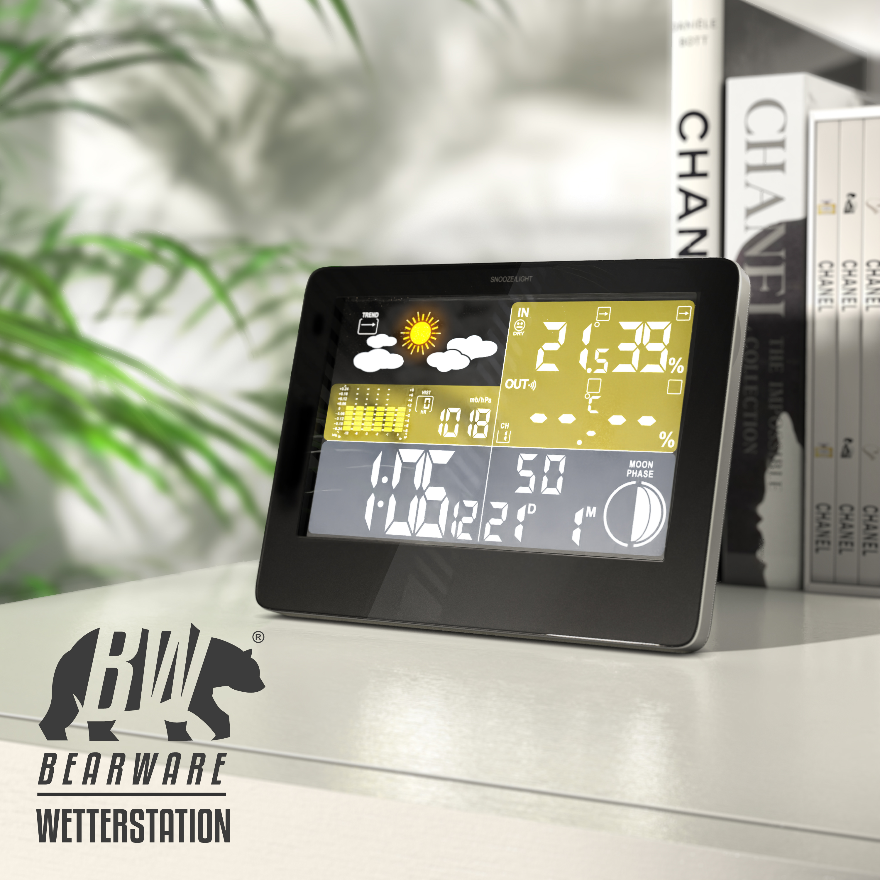 Eine elektronische Wetterstation mit digitalem Display steht auf einem Tisch vor Chanel-Büchern und Pflanzen.
