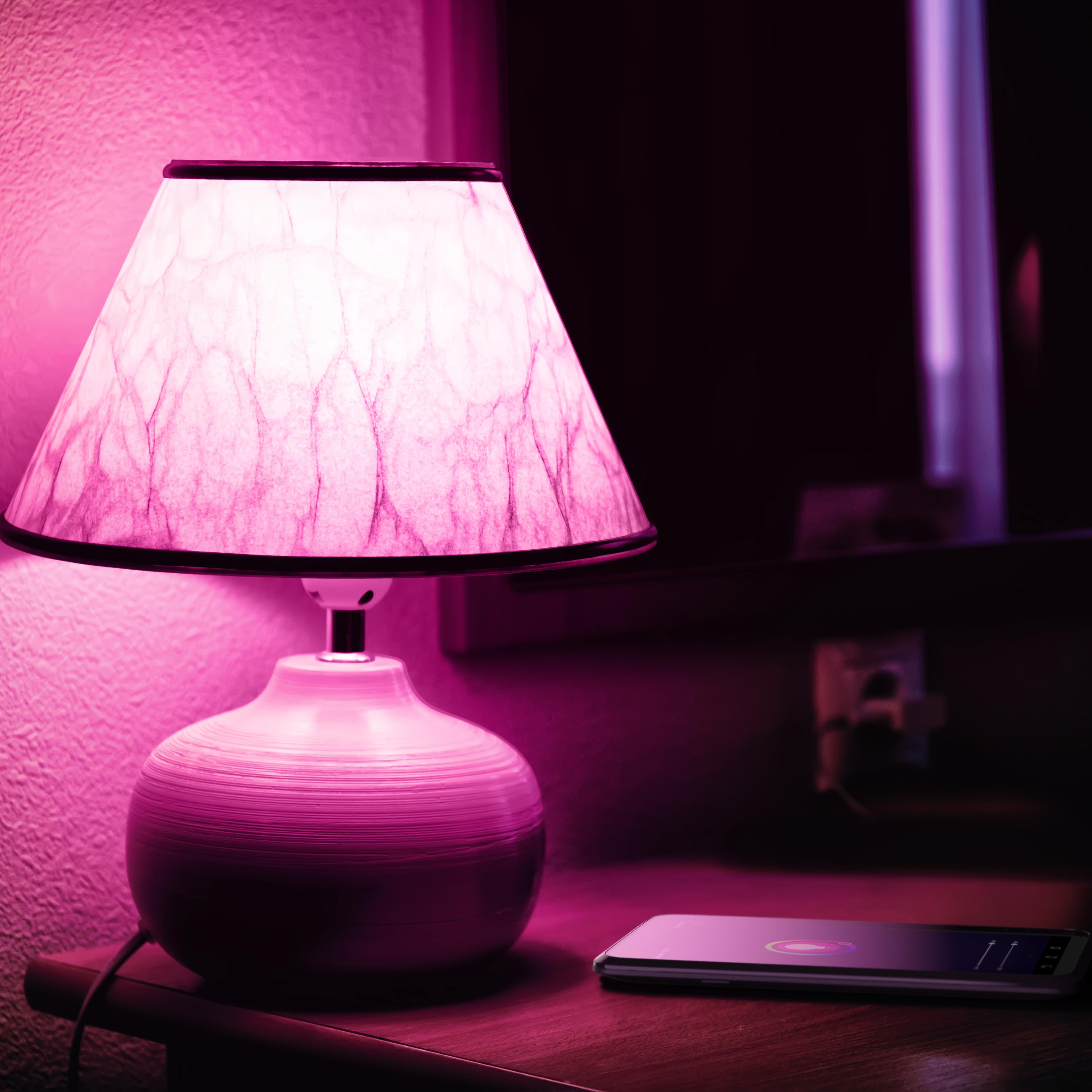 Eine rosafarbene Tischlampe mit gemustertem Schirm und ein Smartphone stehen auf einem Nachttisch.
