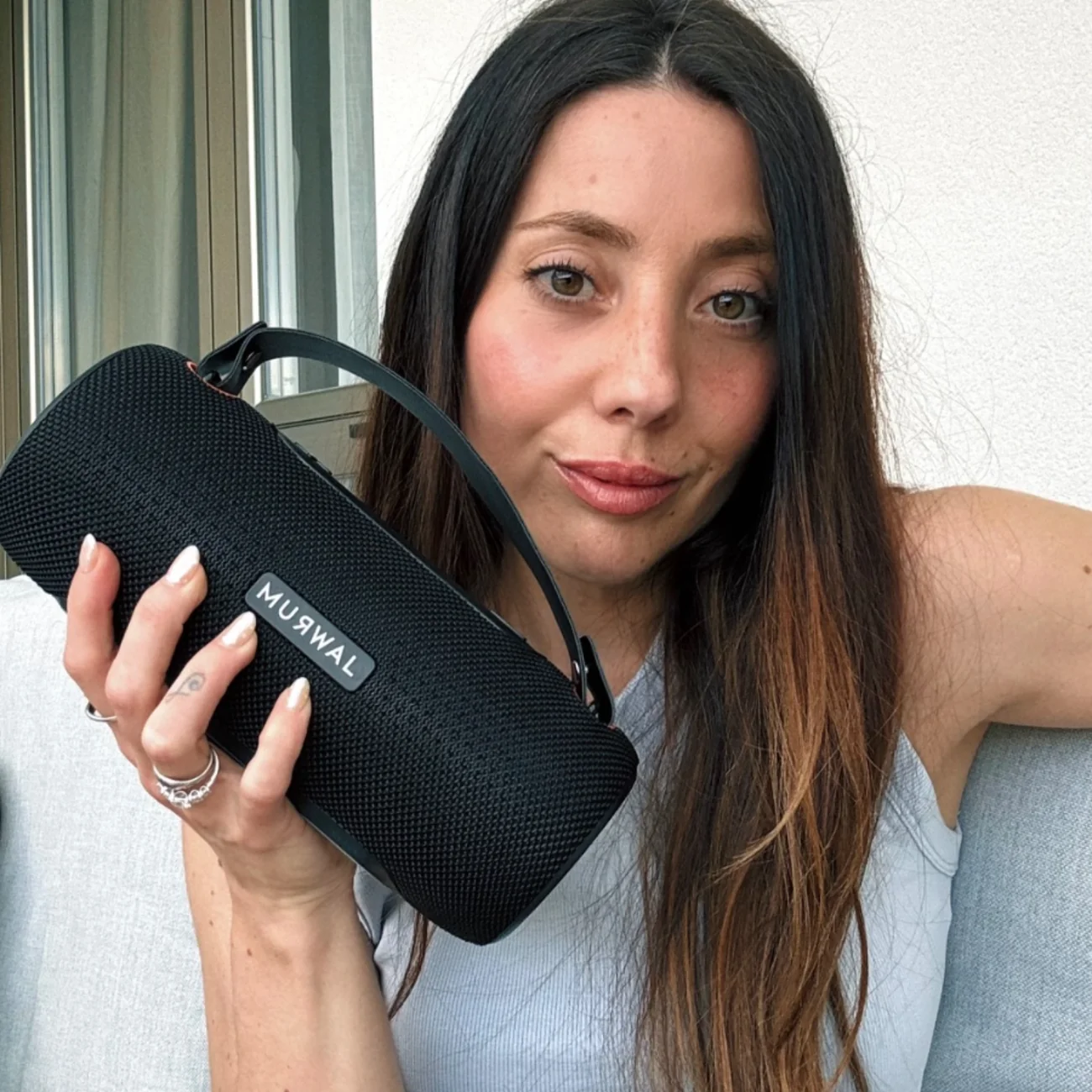 Una mujer con cabello largo y oscuro sostiene un altavoz Bluetooth Muswal negro.