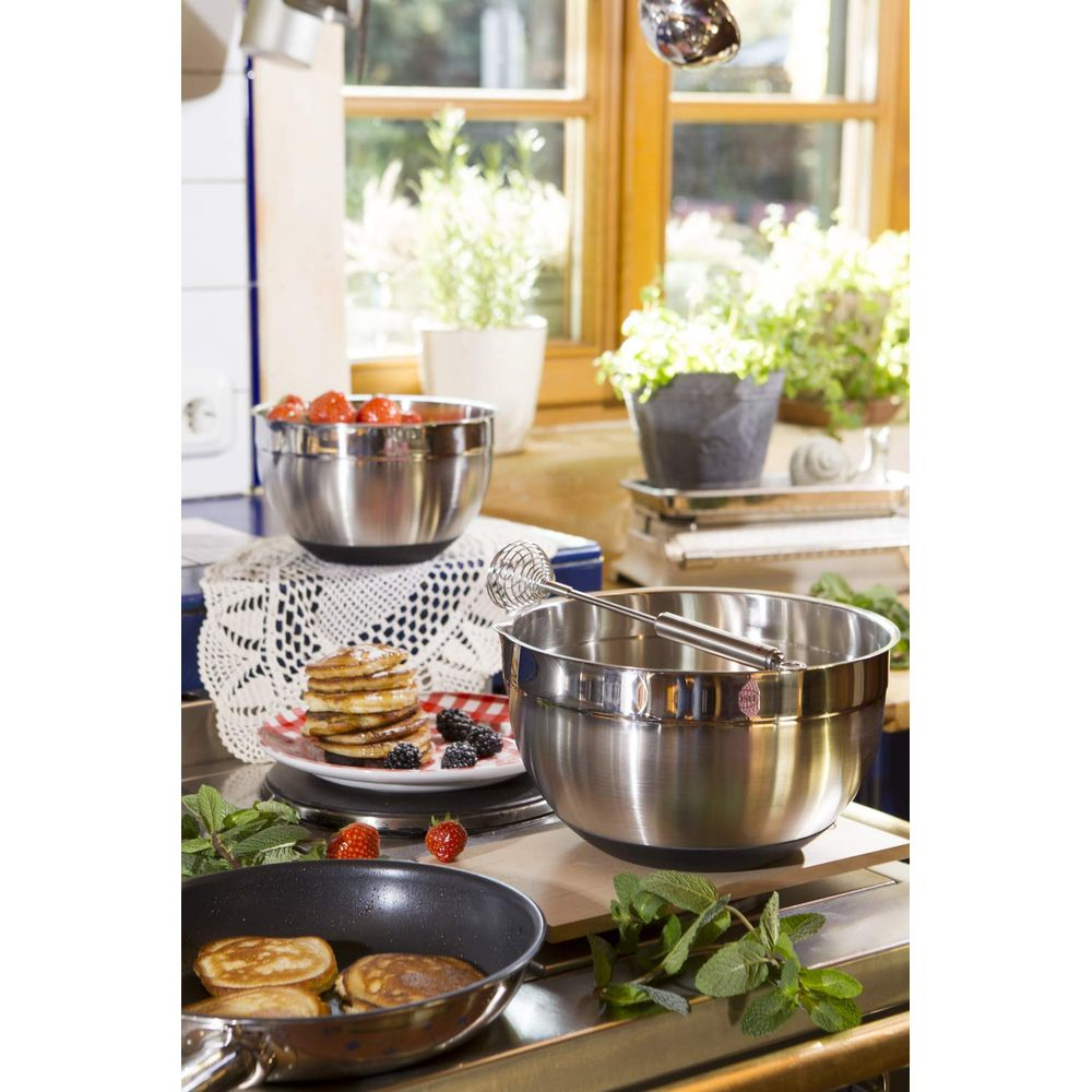 Scena di cucina con ciotole in acciaio inox, pancake su un piatto, fragole fresche ed erbe aromatiche.