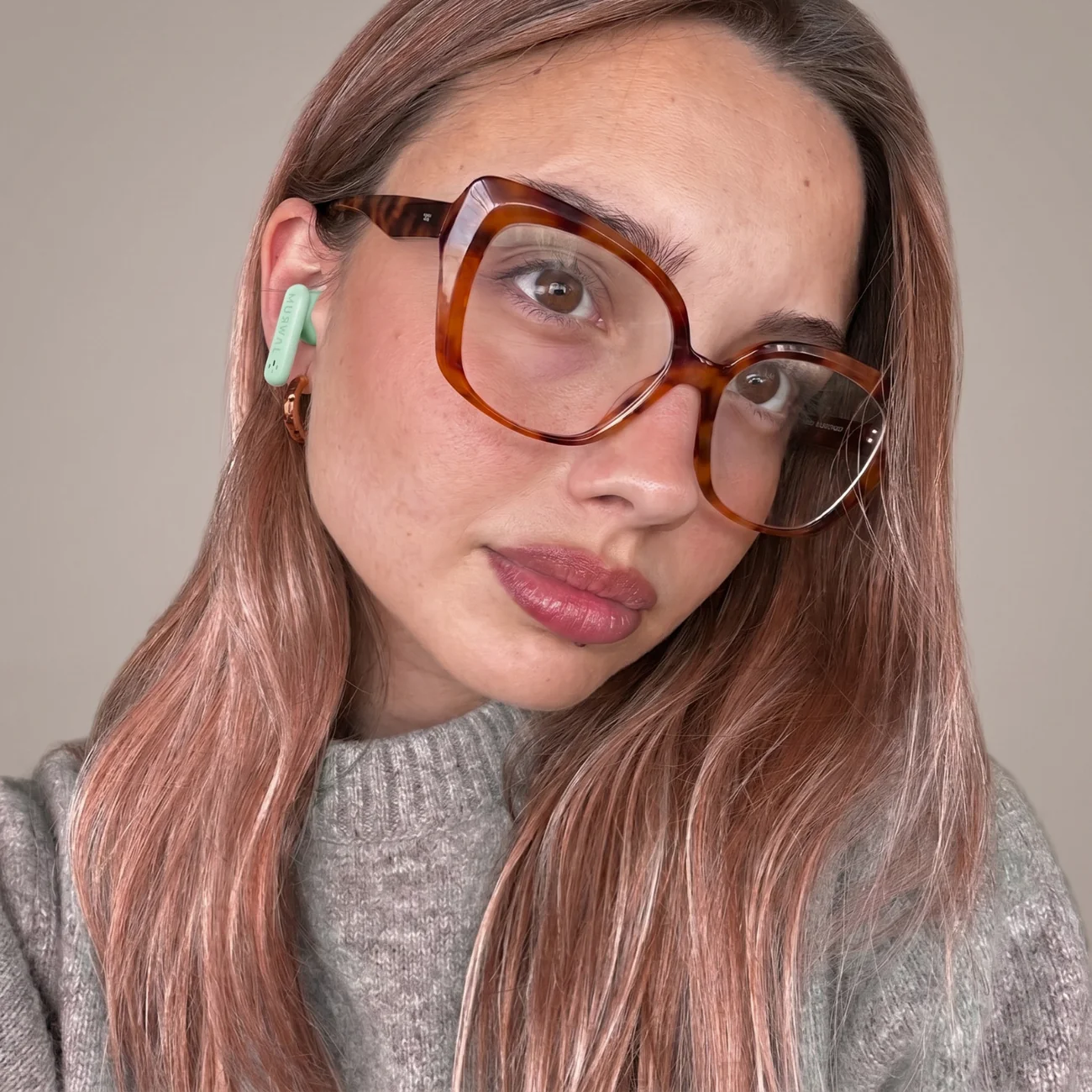 Una mujer con cabello castaño rosado con gafas carey, un suéter gris y auriculares verde menta.