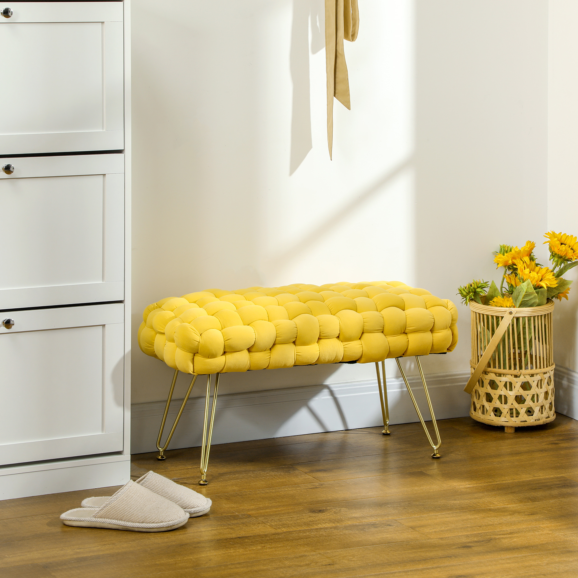 Banco capitoné amarillo con patas doradas, mueble blanco, zapatillas y girasoles en canasta sobre suelo de madera.