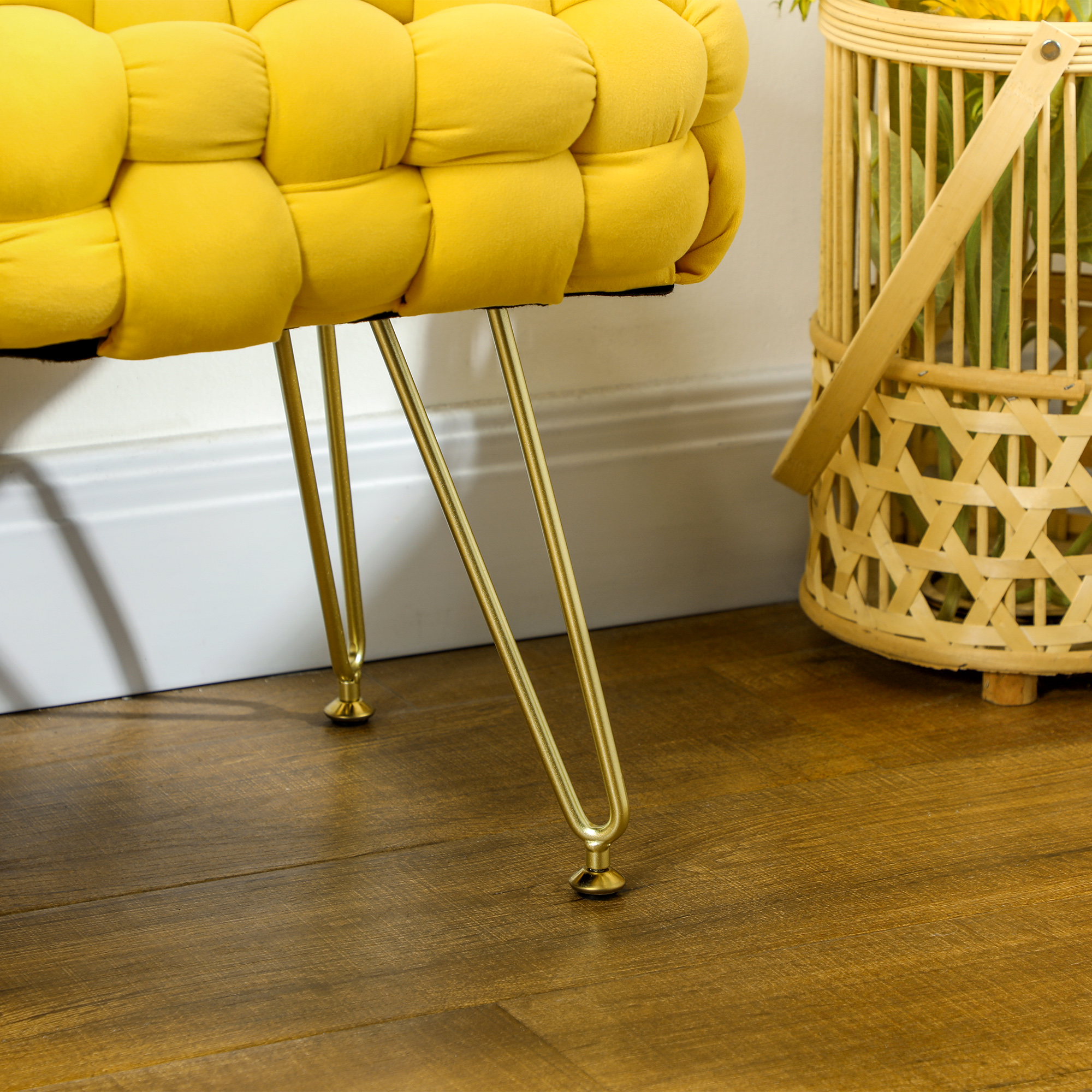 Banco amarillo capitoné con patas de metal dorado en suelo de madera, con cesta de mimbre al lado.