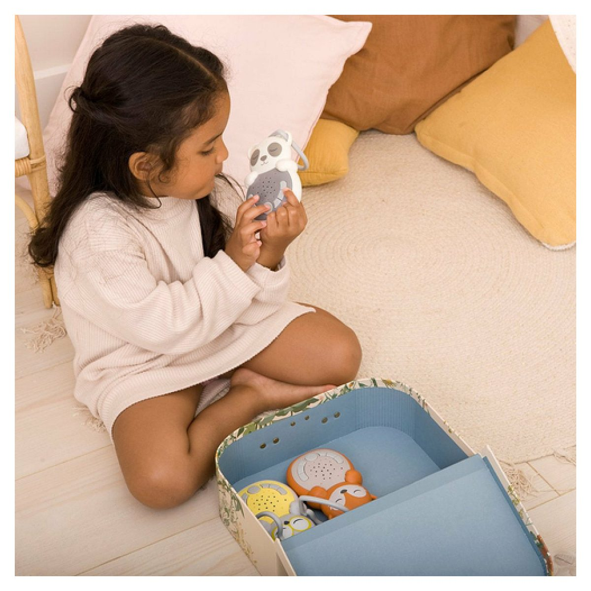 Ein junges Mädchen sitzt auf einem Teppich und hält ein kleines Panda-Spielzeug, während andere Spielzeuge in einer Kiste liegen.