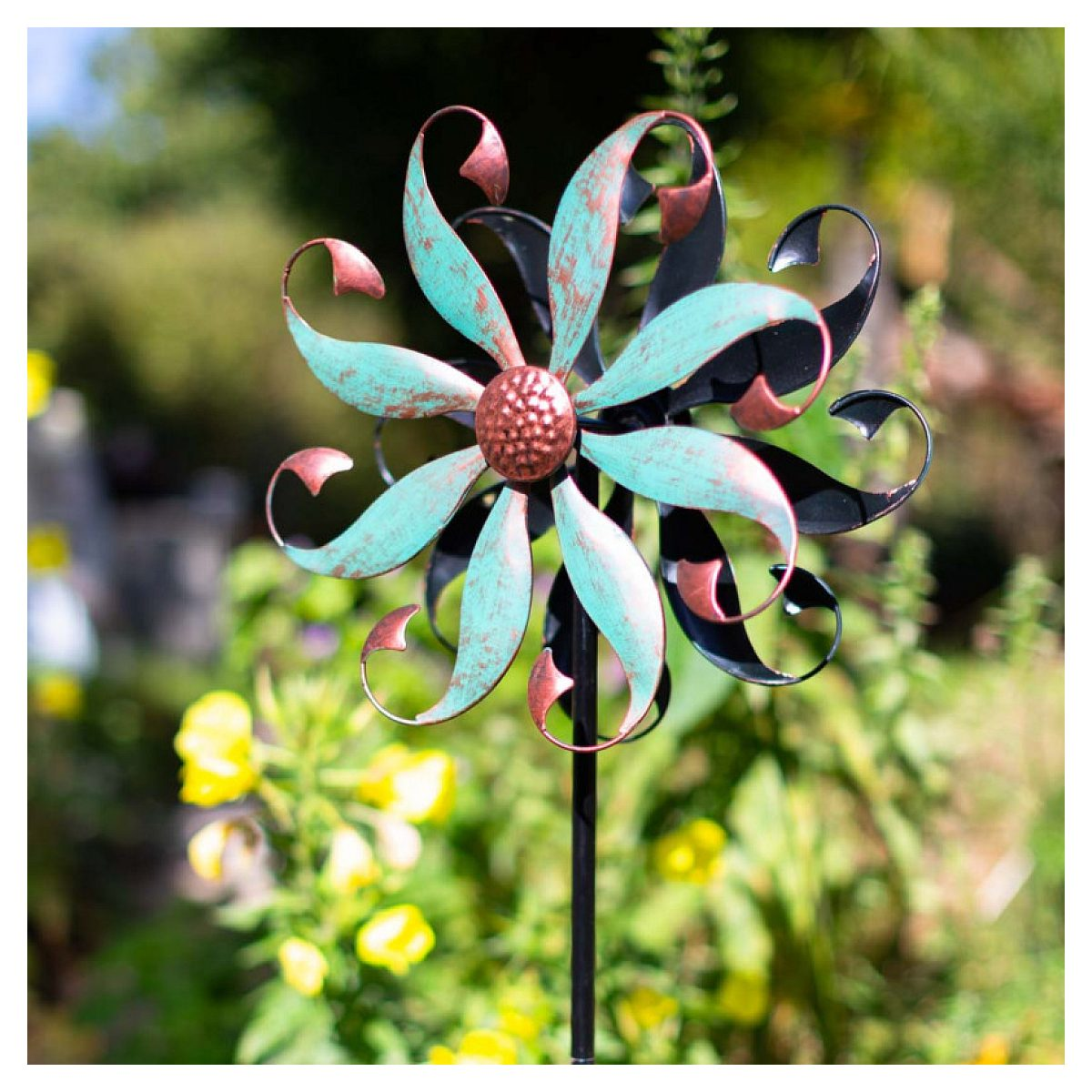 Ein rot-türkisfarbenes Windspiel aus Metall steht auf einem Stab in einem sonnigen, grünen Garten mit gelben Blumen.