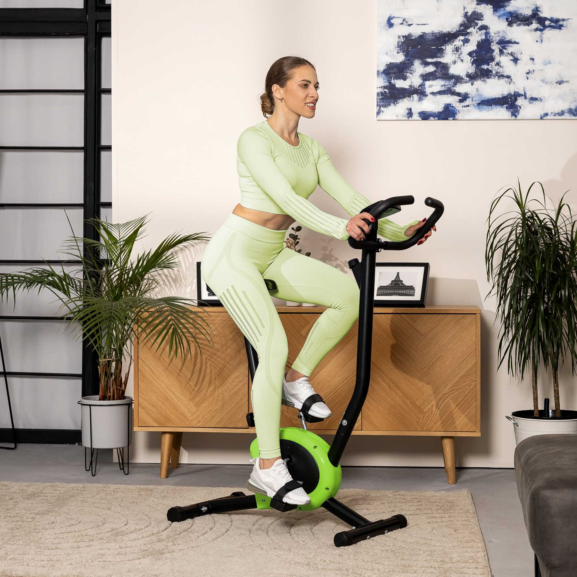 Eine Frau in hellgrüner Sportkleidung trainiert zu Hause auf einem grünen Heimtrainer.