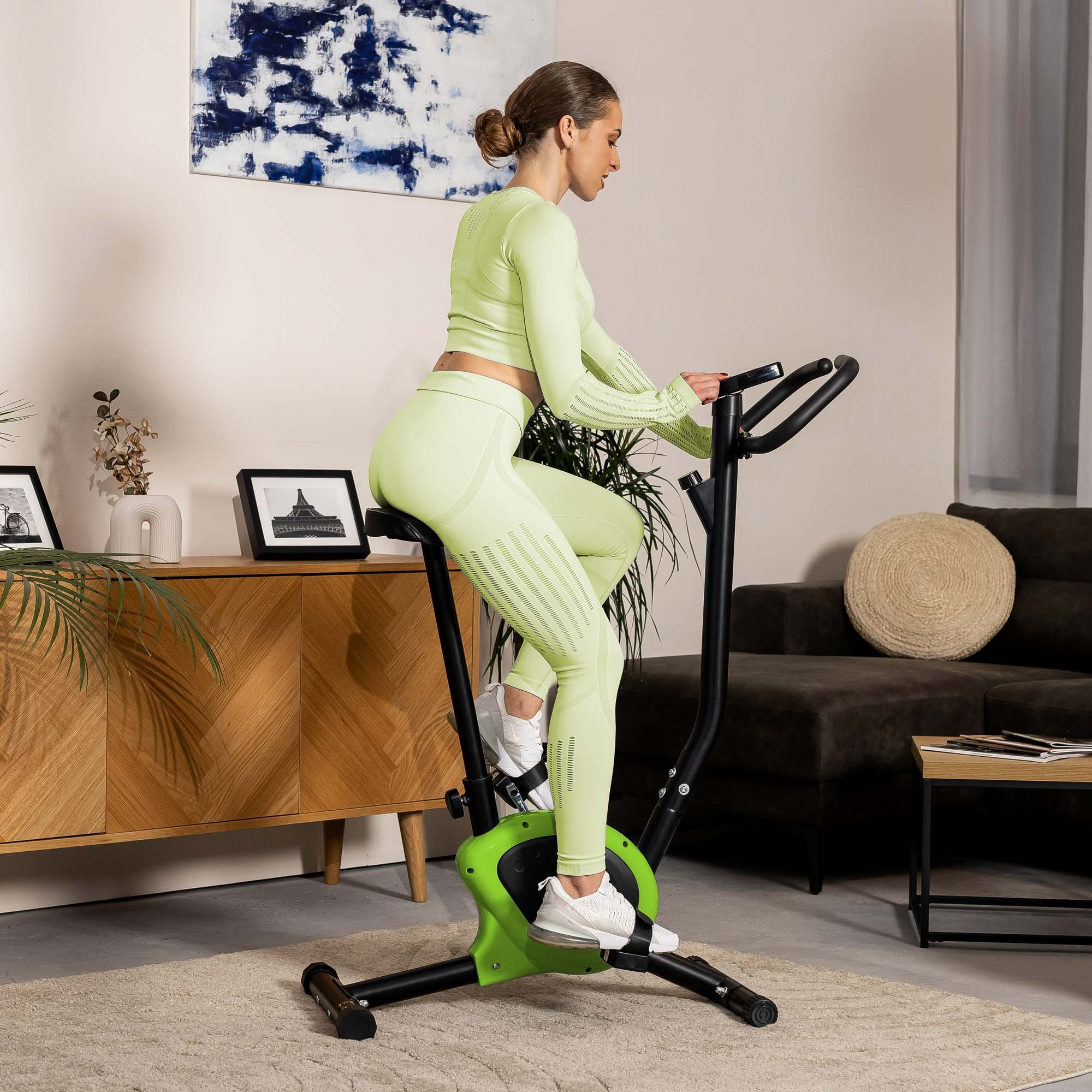 Eine Frau in limettengrüner Sportkleidung trainiert zu Hause auf einem schwarzen Heimtrainer.