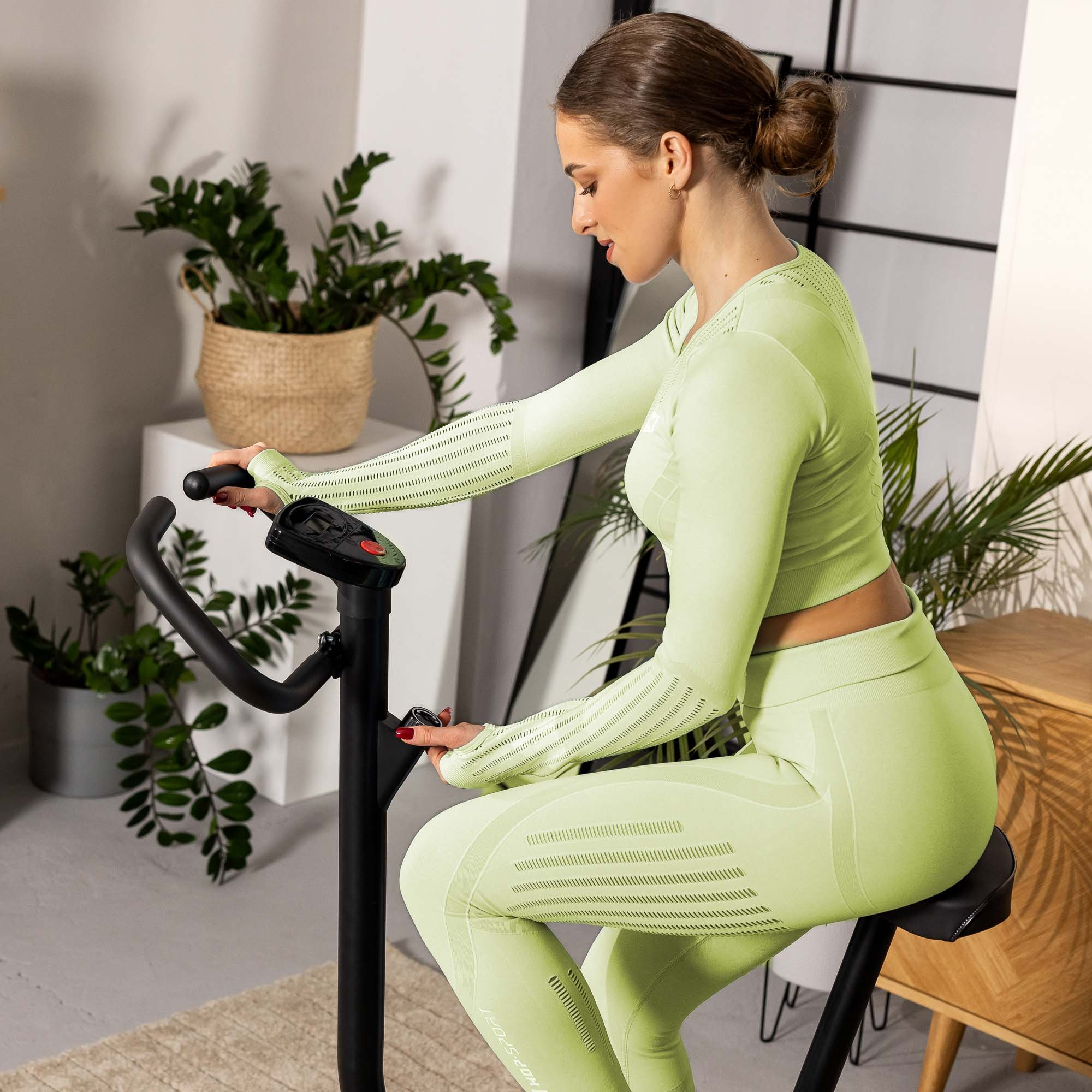 Eine Frau in hellgrüner Sportbekleidung sitzt auf einem schwarzen Heimtrainer in einem hellen Raum mit Pflanzen.