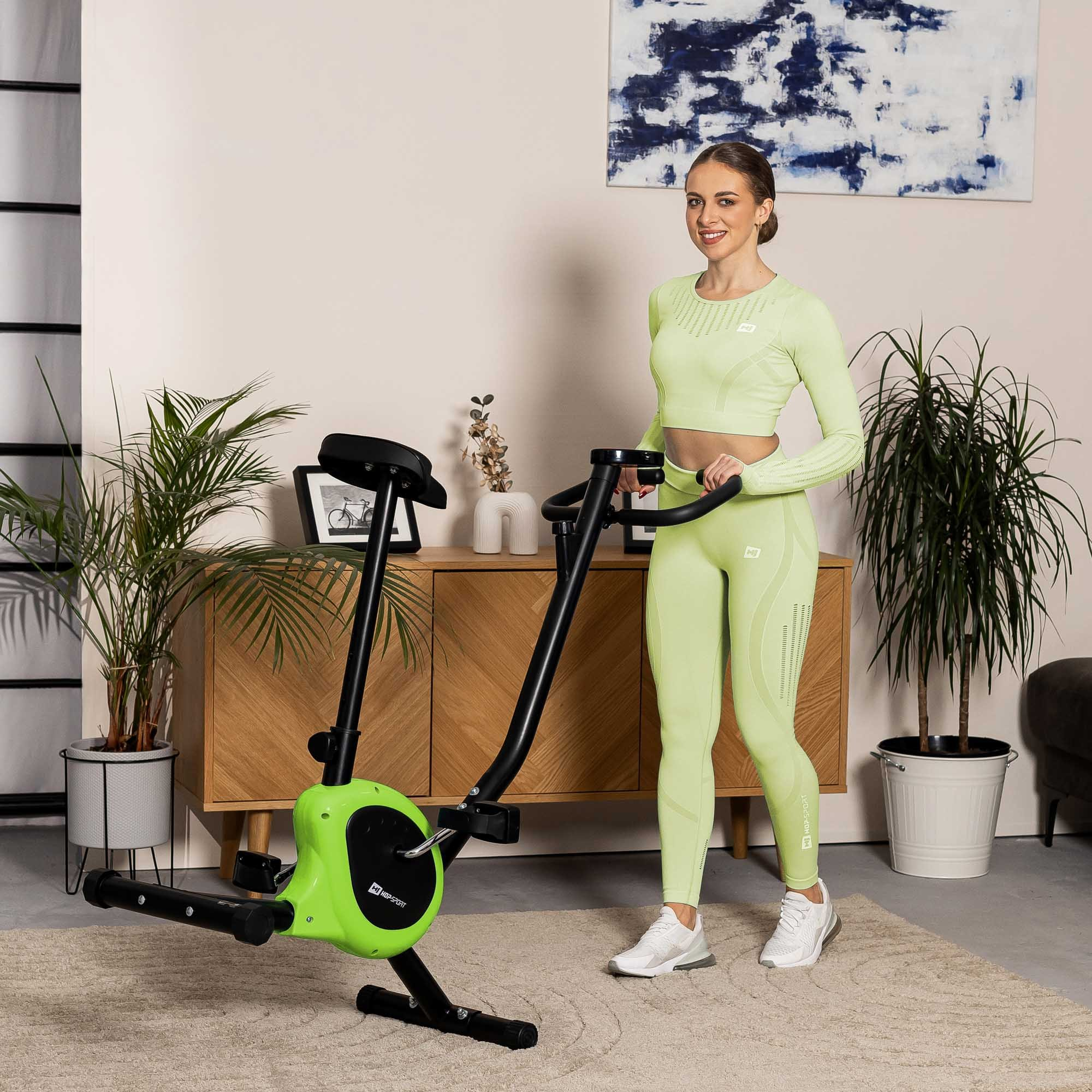 Eine Frau in hellgrüner Sportkleidung steht neben einem Heimtrainer in einem hellen Zimmer mit Topfpflanzen.