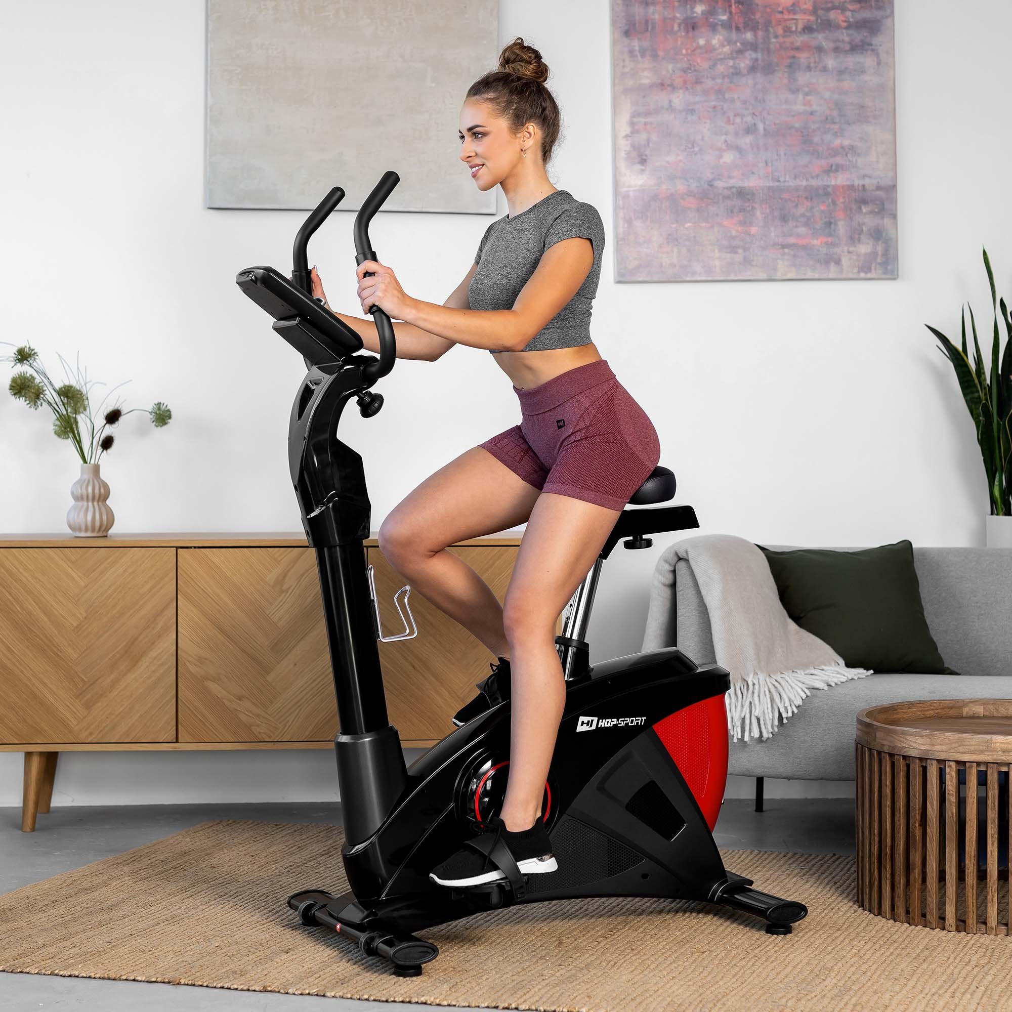 Eine Frau trainiert auf einem schwarz-roten Heimtrainer in einem hellen Wohnzimmer.
