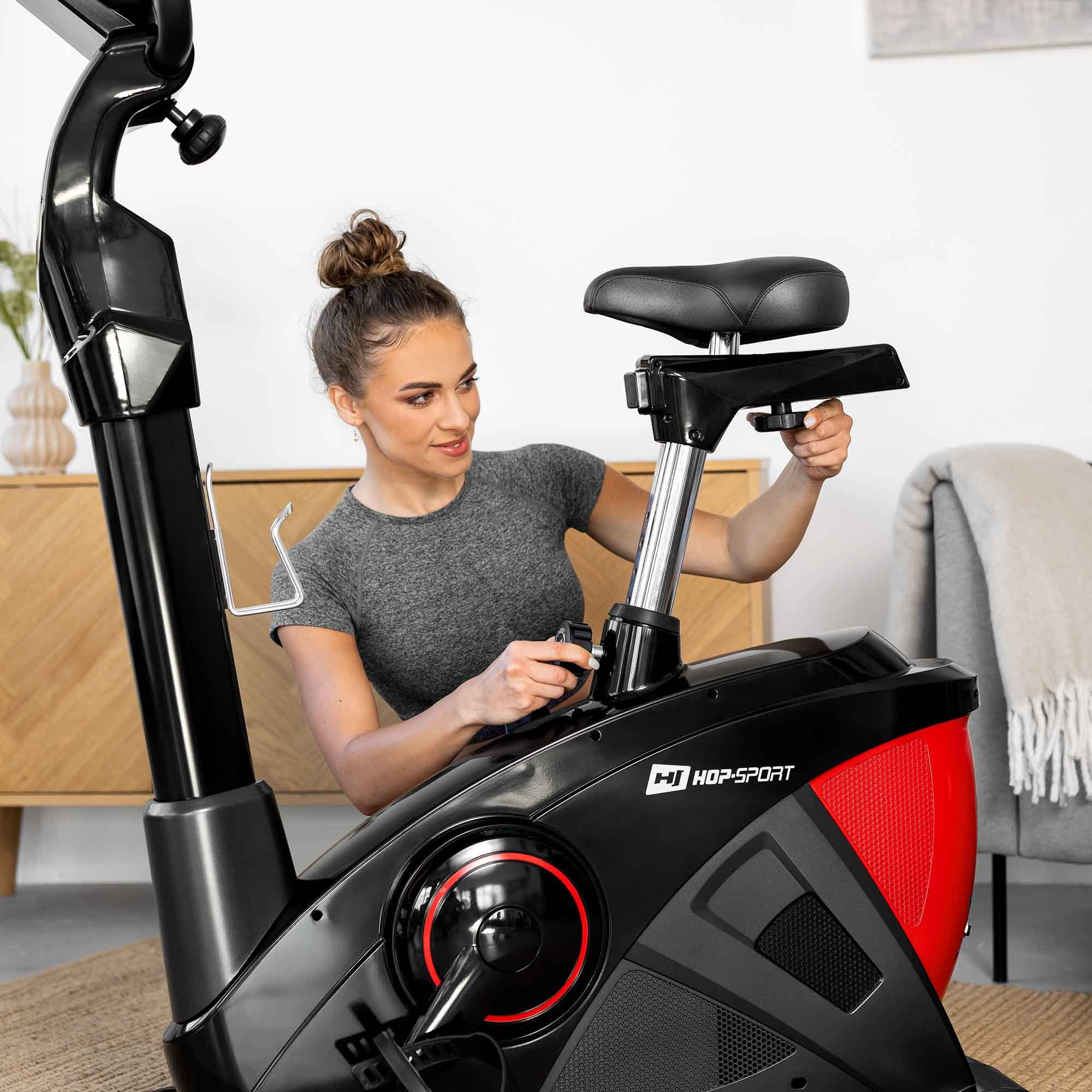 Eine Frau stellt den Sattel und den Widerstandsknopf an einem schwarz-roten Heimtrainer von Hop-Sport ein.