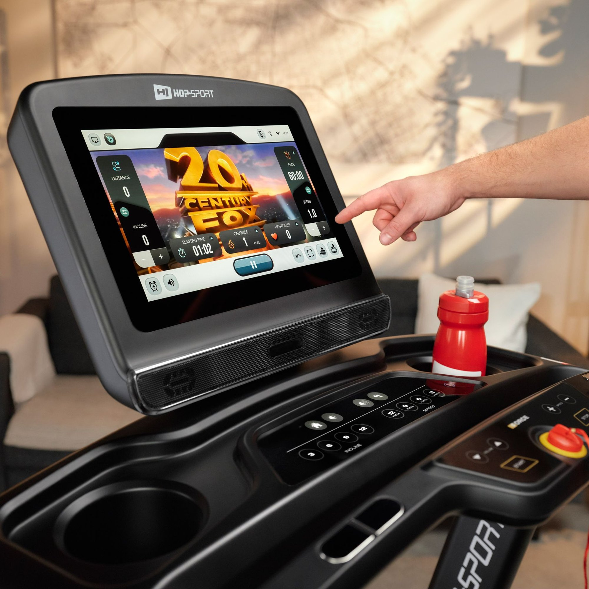 Eine Person berührt den Touchscreen eines Hop-Sport Laufbands mit Videowiedergabe, daneben eine rote Trinkflasche.