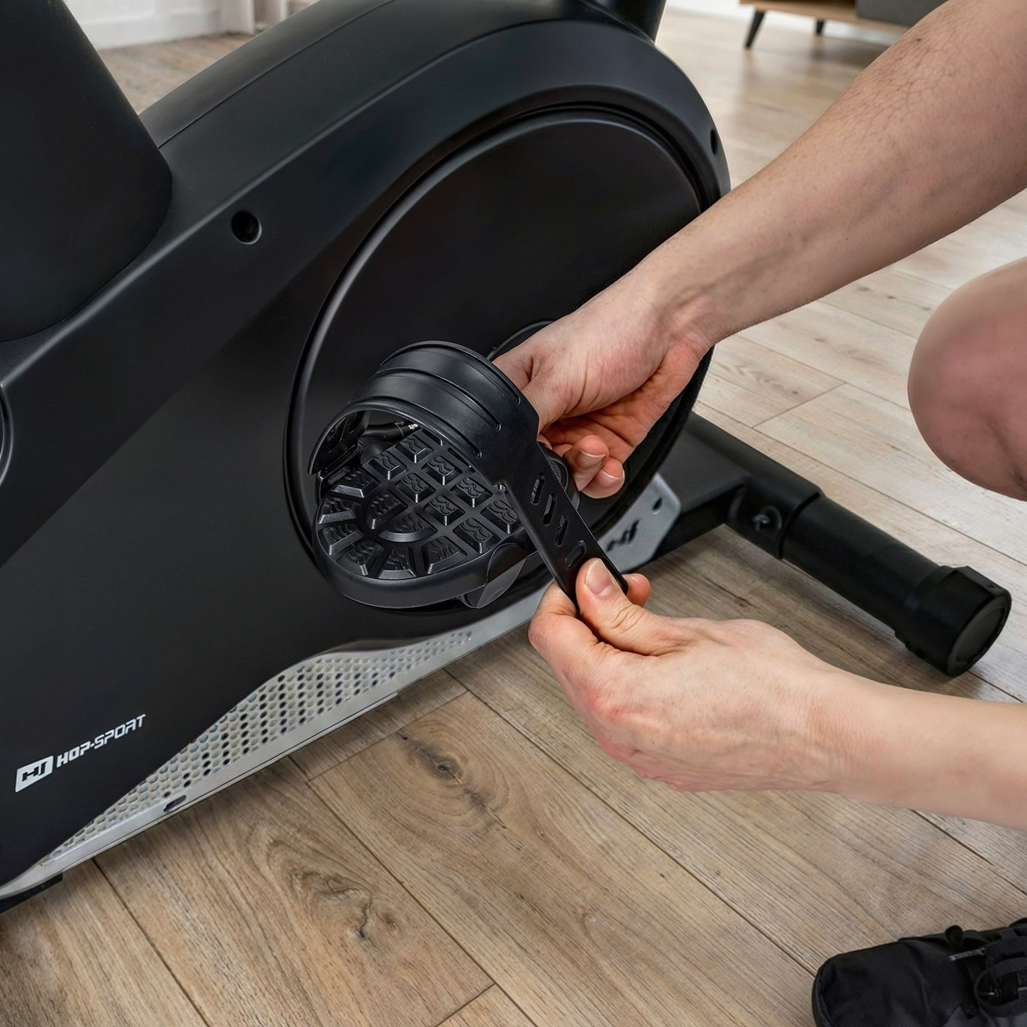 Eine Person stellt den schwarzen Riemen am Pedal eines Heimtrainers auf einem Holzboden ein.