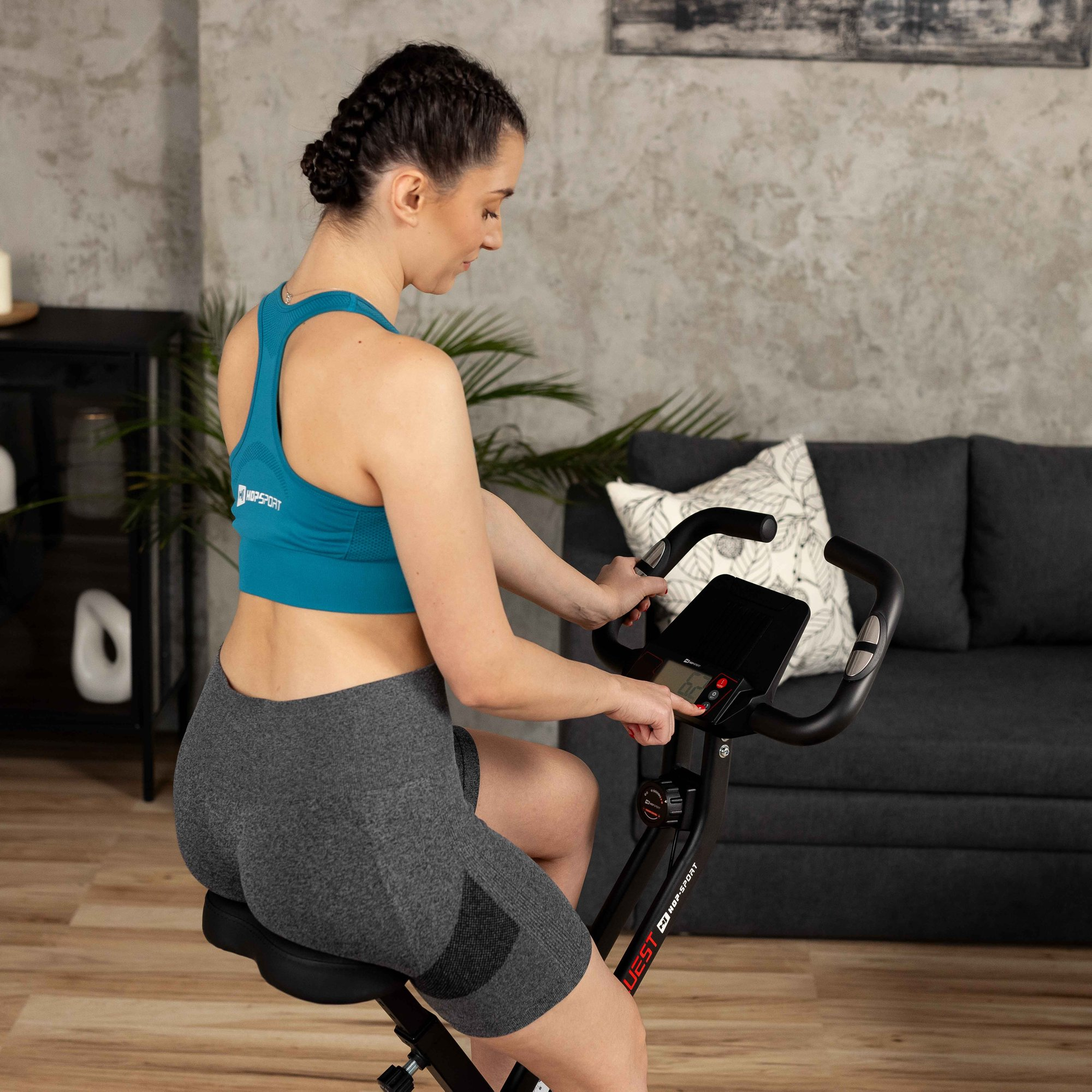 Eine Frau im blauen Sporttop und grauen Shorts trainiert auf einem Heimtrainer in einem Wohnzimmer.