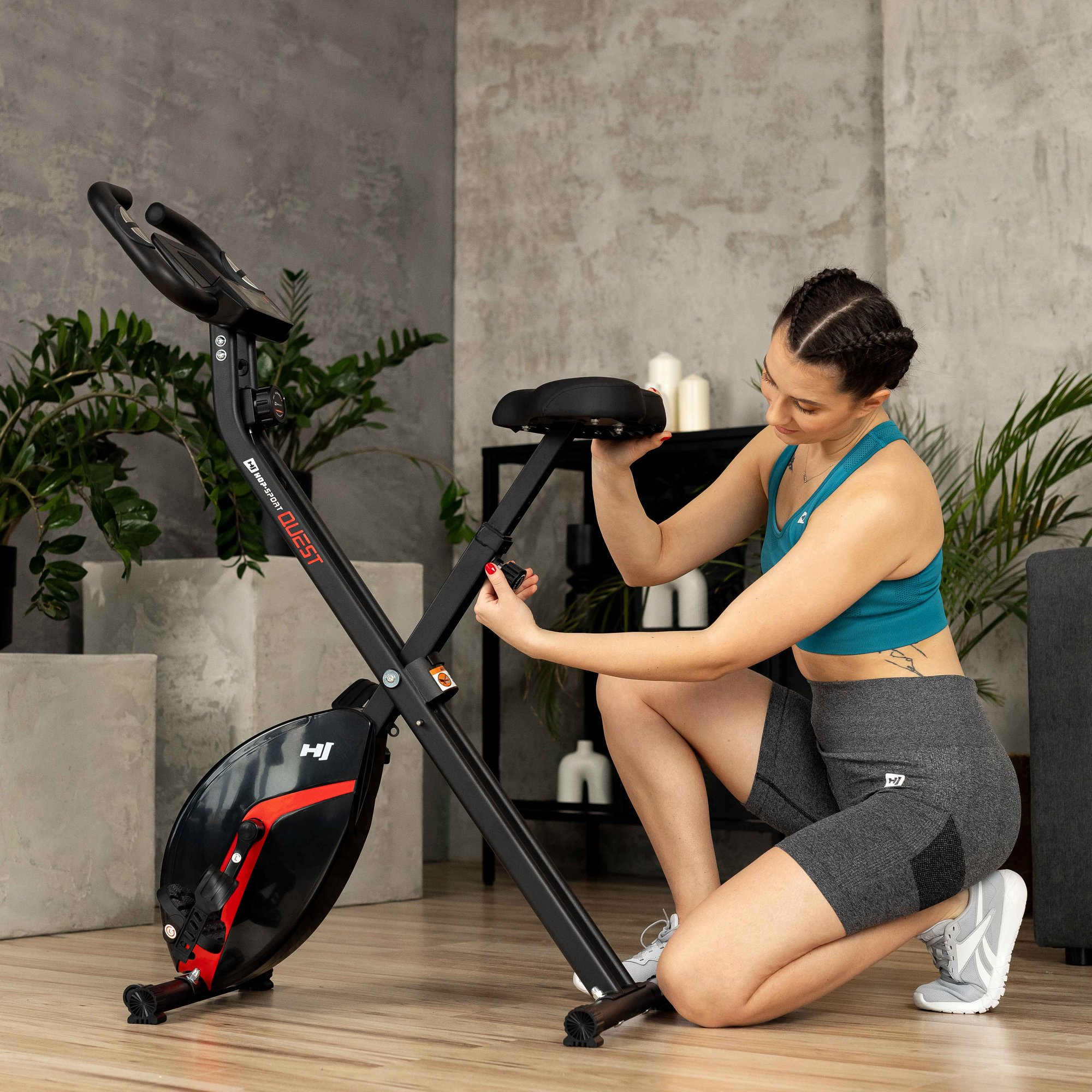 Eine Frau in Sportkleidung kniet und stellt die Sattelhöhe eines schwarzen Heimtrainers ein.